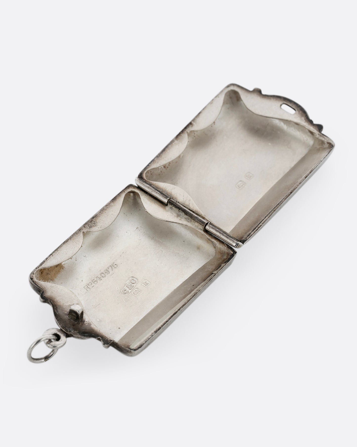 A silver rectangular locket with a filigreed top and hinged bottom. Shown open lying flat from the top.