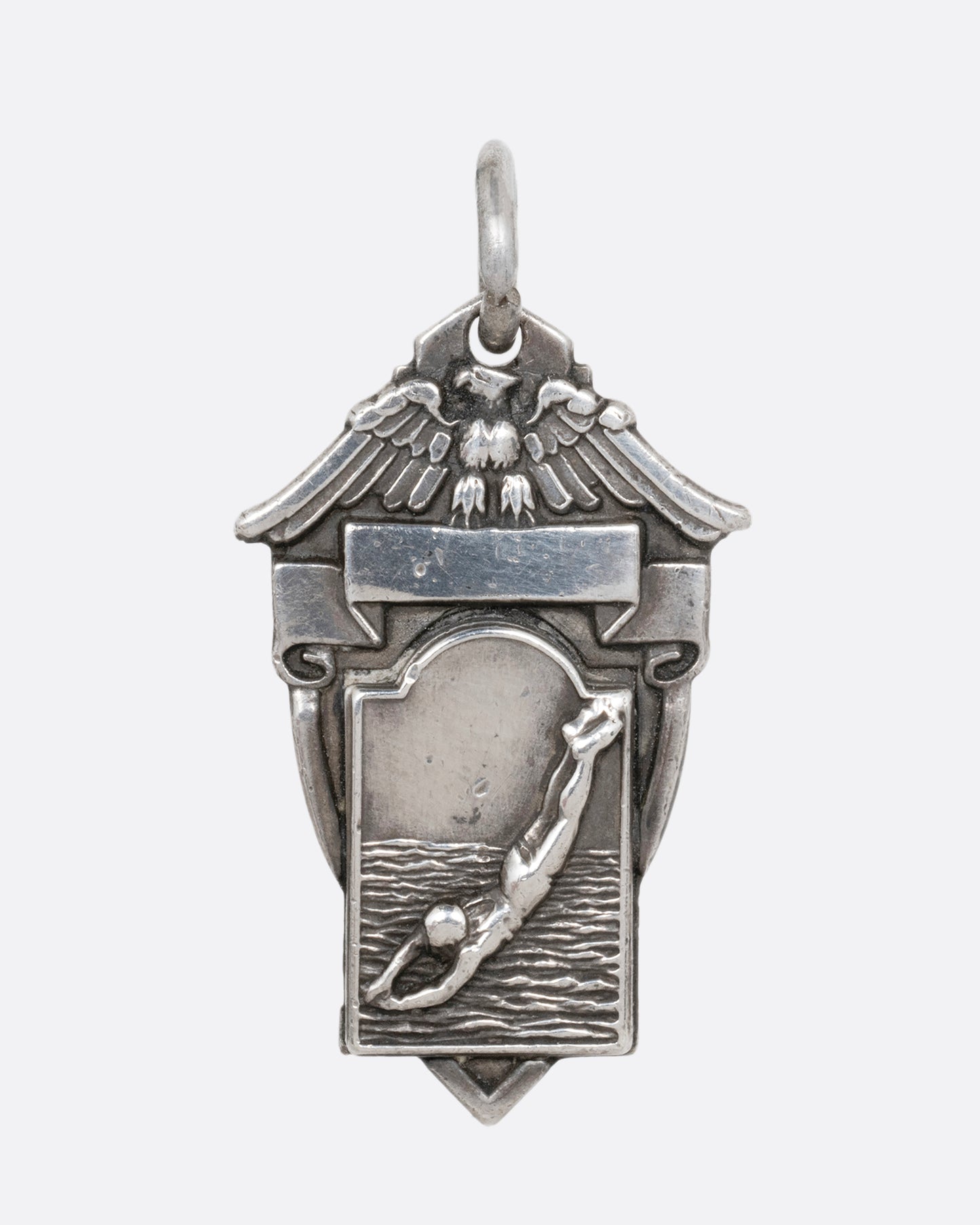 A silver crest shaped pendant with a figure diving and an eagle sitting atop a ribbon. Shown from the front.
