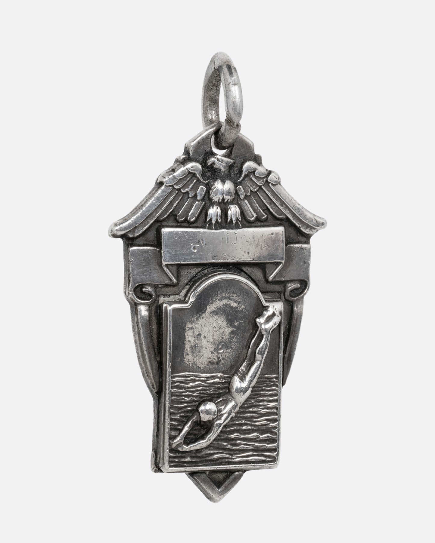 A silver crest shaped pendant with a figure diving and an eagle sitting atop a ribbon. Shown from the side.