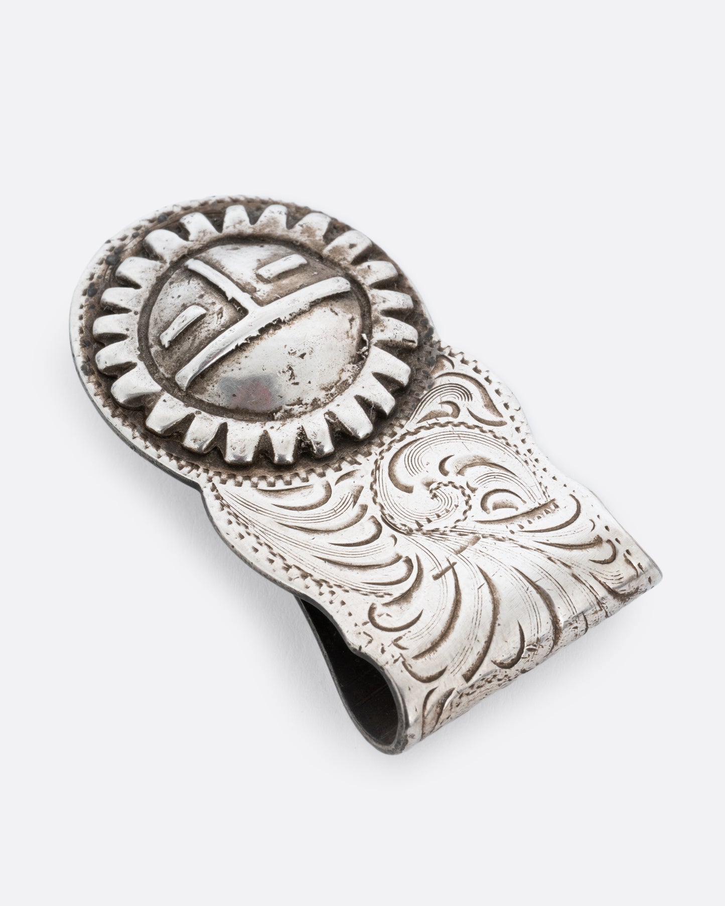 A silver money clip with a swirling engraved pattern and the Hopi sun spirit and creator, Tawa, on the end of the front. Shown lying flat from the top.