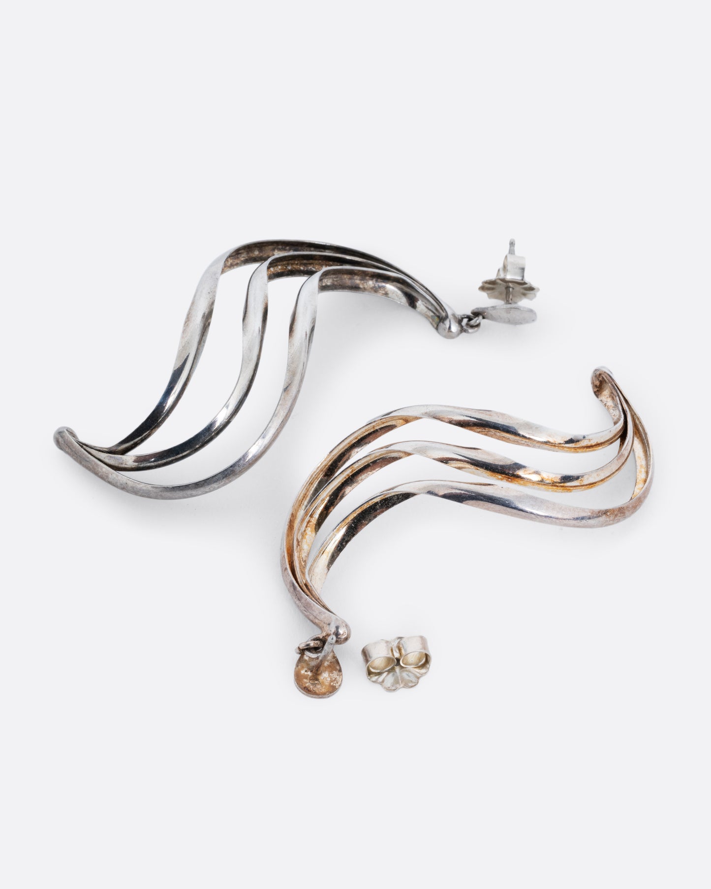 Silver dangle earrings with stud tops and three layers of silver ribbons in an S shape. One earring is shown lying flat from the front. The other earring is shown lying flat open from the back.