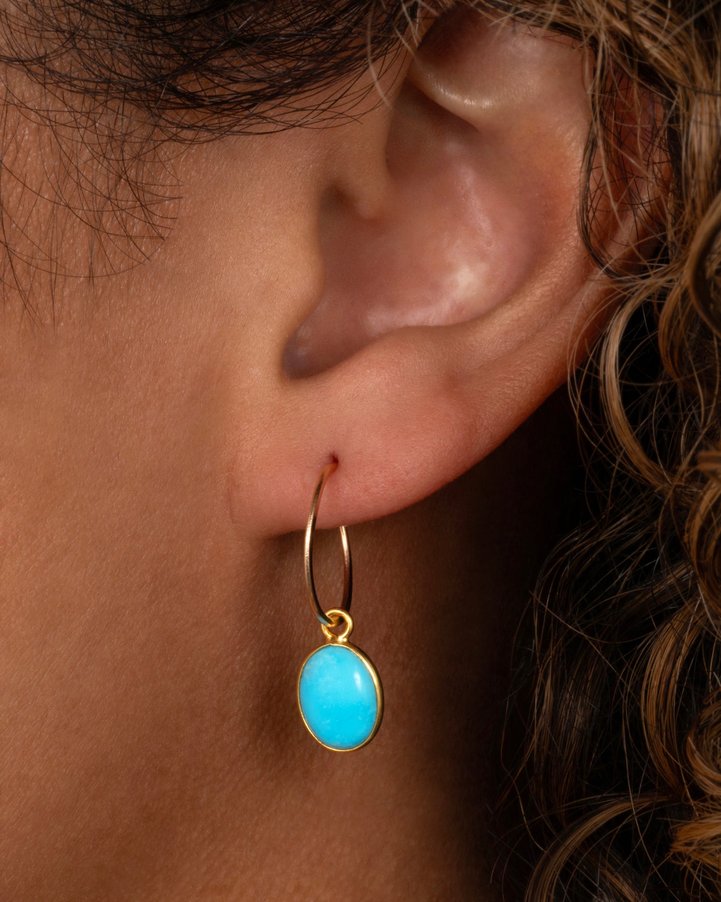 A Sleeping Beauty Turquoise dangle set in yellow gold with a yellow gold bail. Shown on a yellow gold hoop from the front on a person.