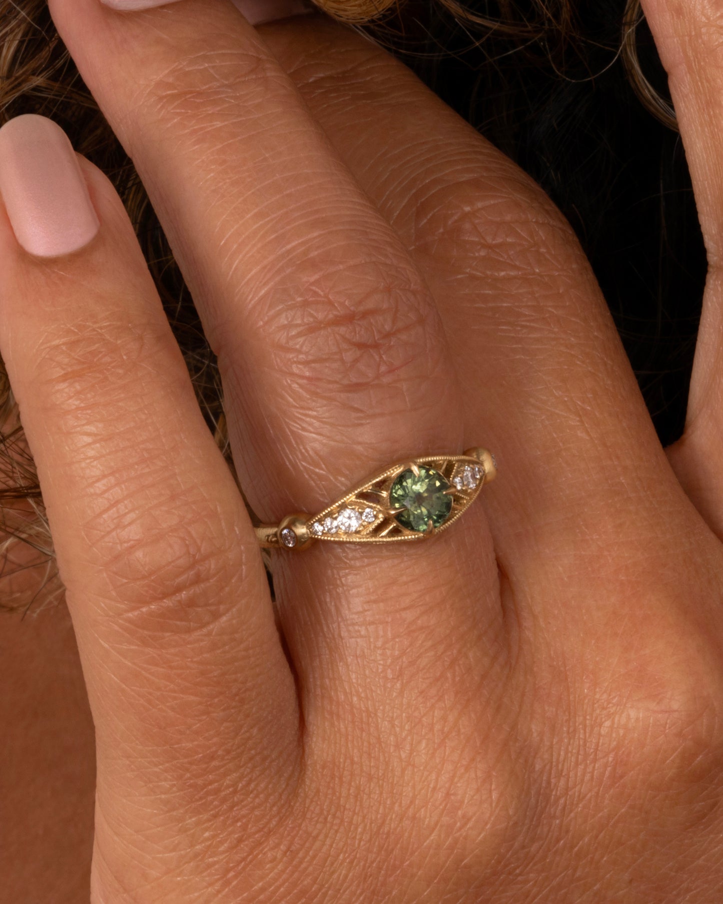 A yellow gold ring with a green Montana sapphire at the center and cutout details and white diamonds. Shown worn on a hand.