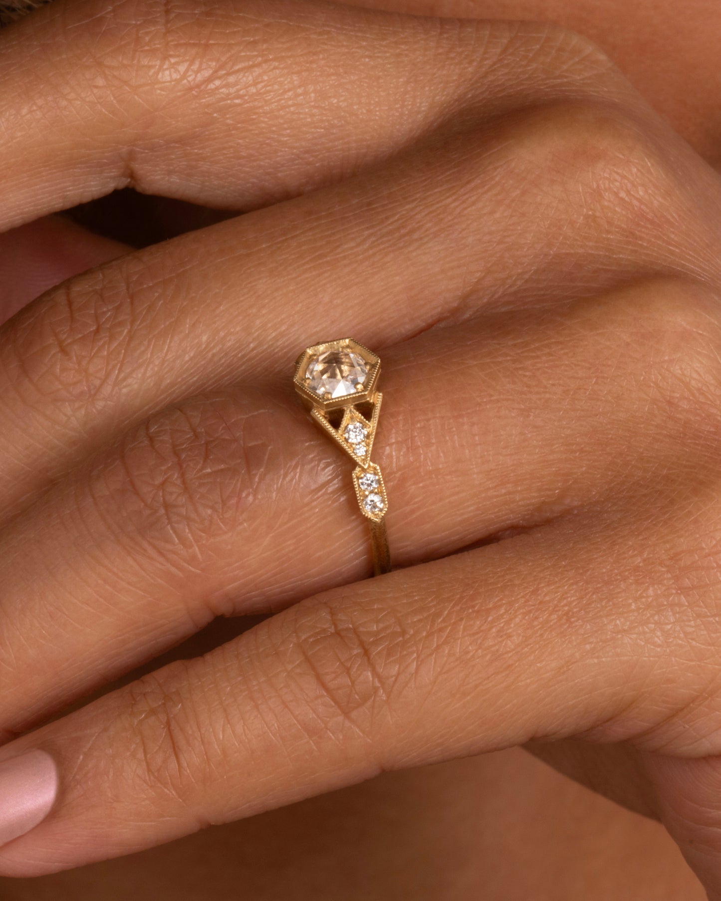 A yellow gold ring with a center diamond and angular side cutouts and diamond accents. Shown from the side on a hand.