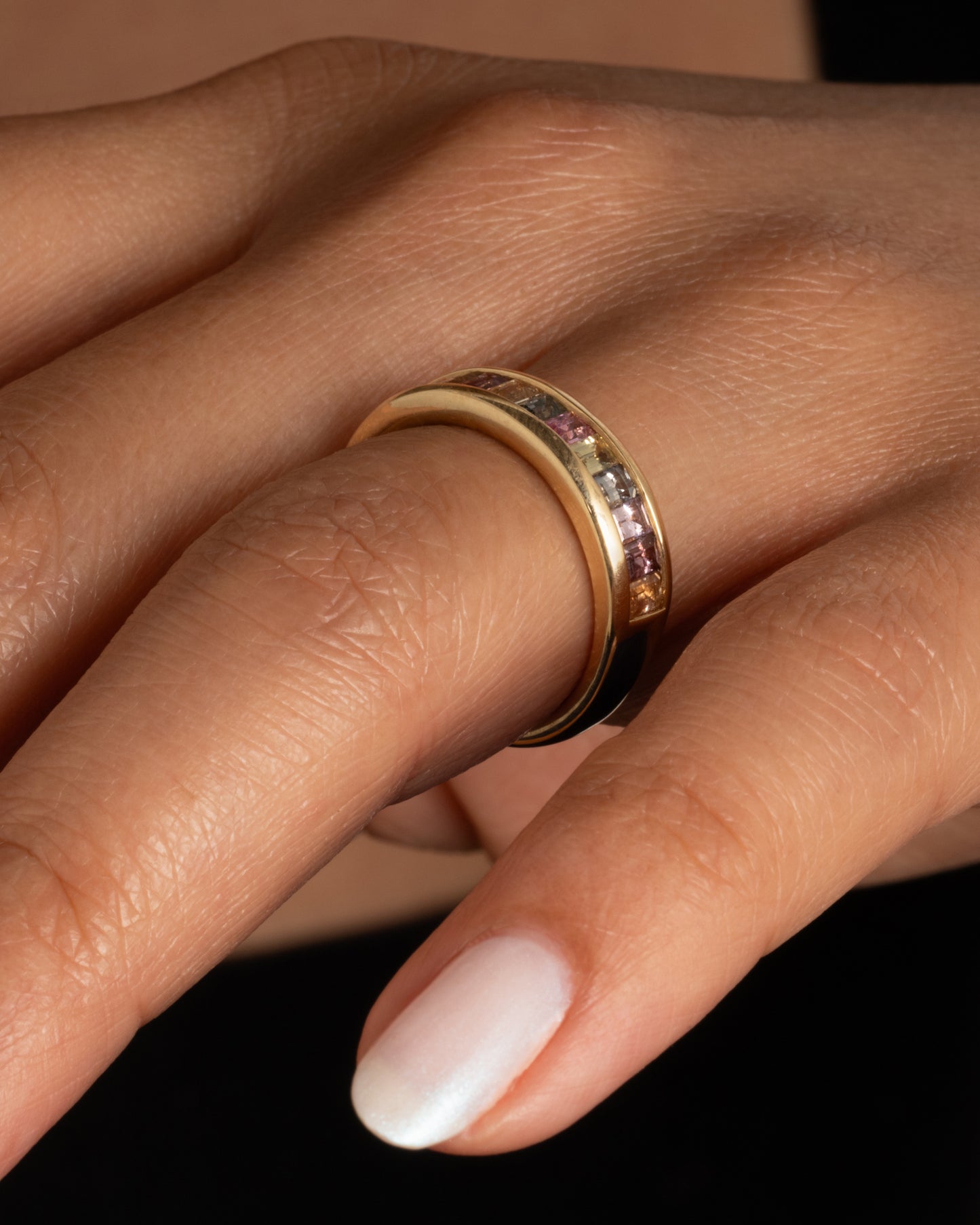 A yellow gold ring with channel set multicolored sapphires. Shown on a hand.