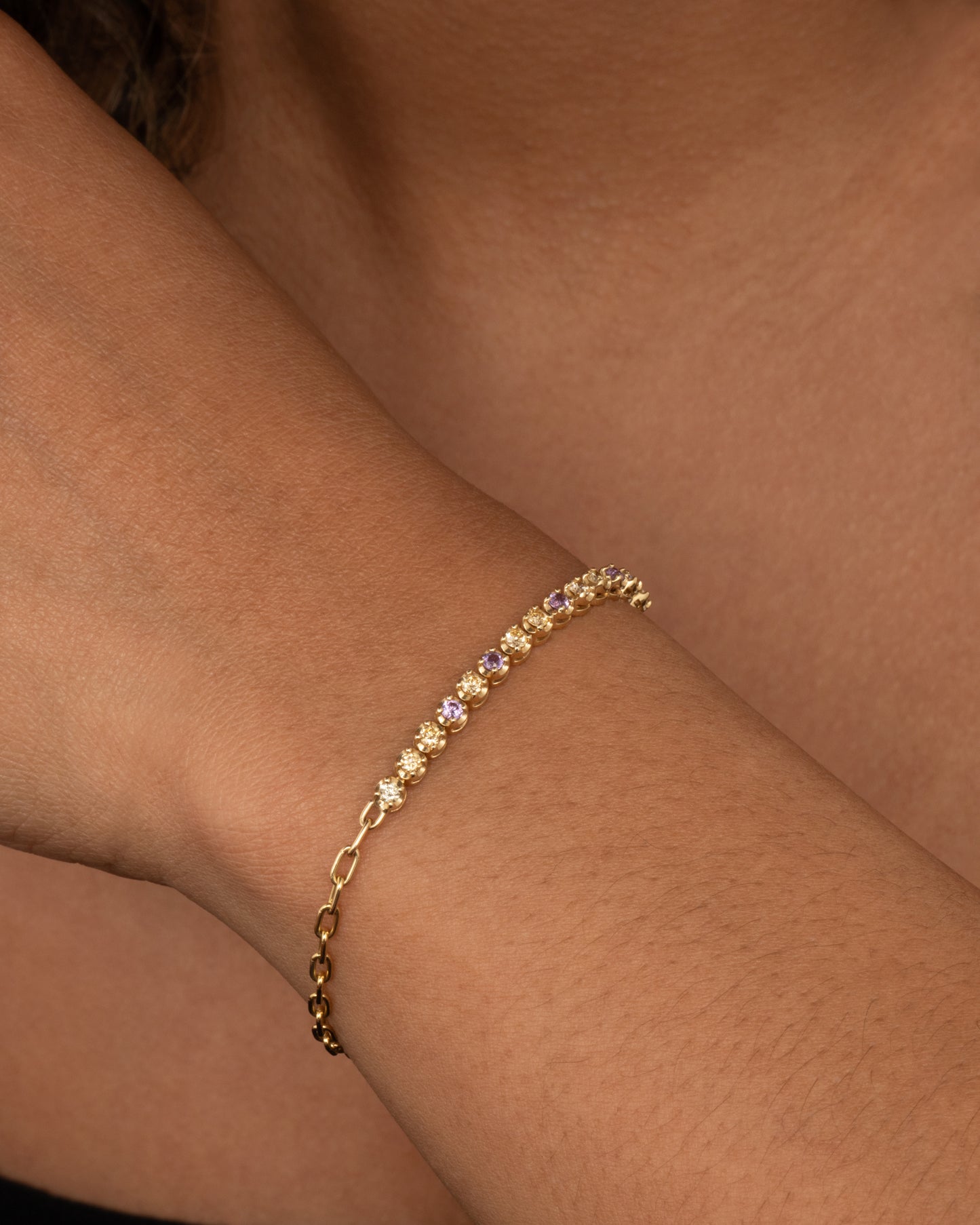 A yellow gold partial tennis bracelet with peach and yellow diamonds and purple sapphires. Shown from the side on a person.