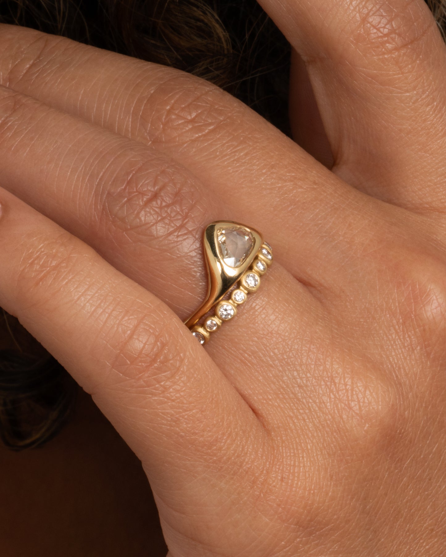 A yellow gold ring with a triangular rose cut diamond. Shown on a finger from the front stacked on top of a diamond eternity band.
