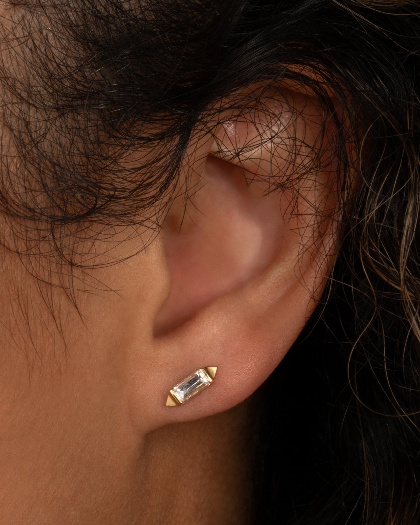 A yellow gold stud earring with a baguette diamond, bookended with points. Shown on an ear.