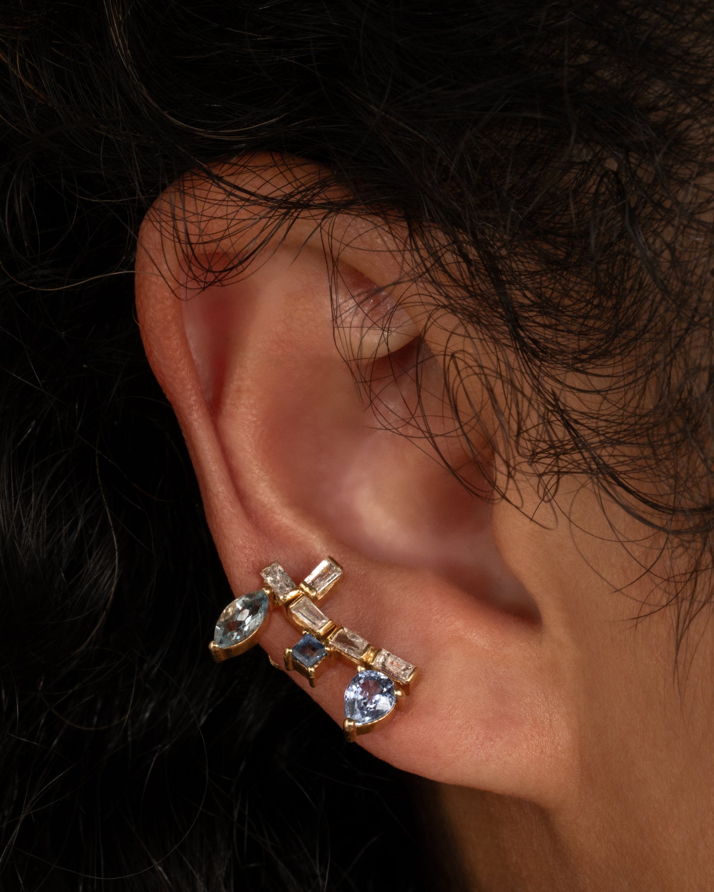 A yellow gold stud earring shaped like a botanical vine made of sapphire, apatite, and diamonds. Shown worn in a second ear piercing.