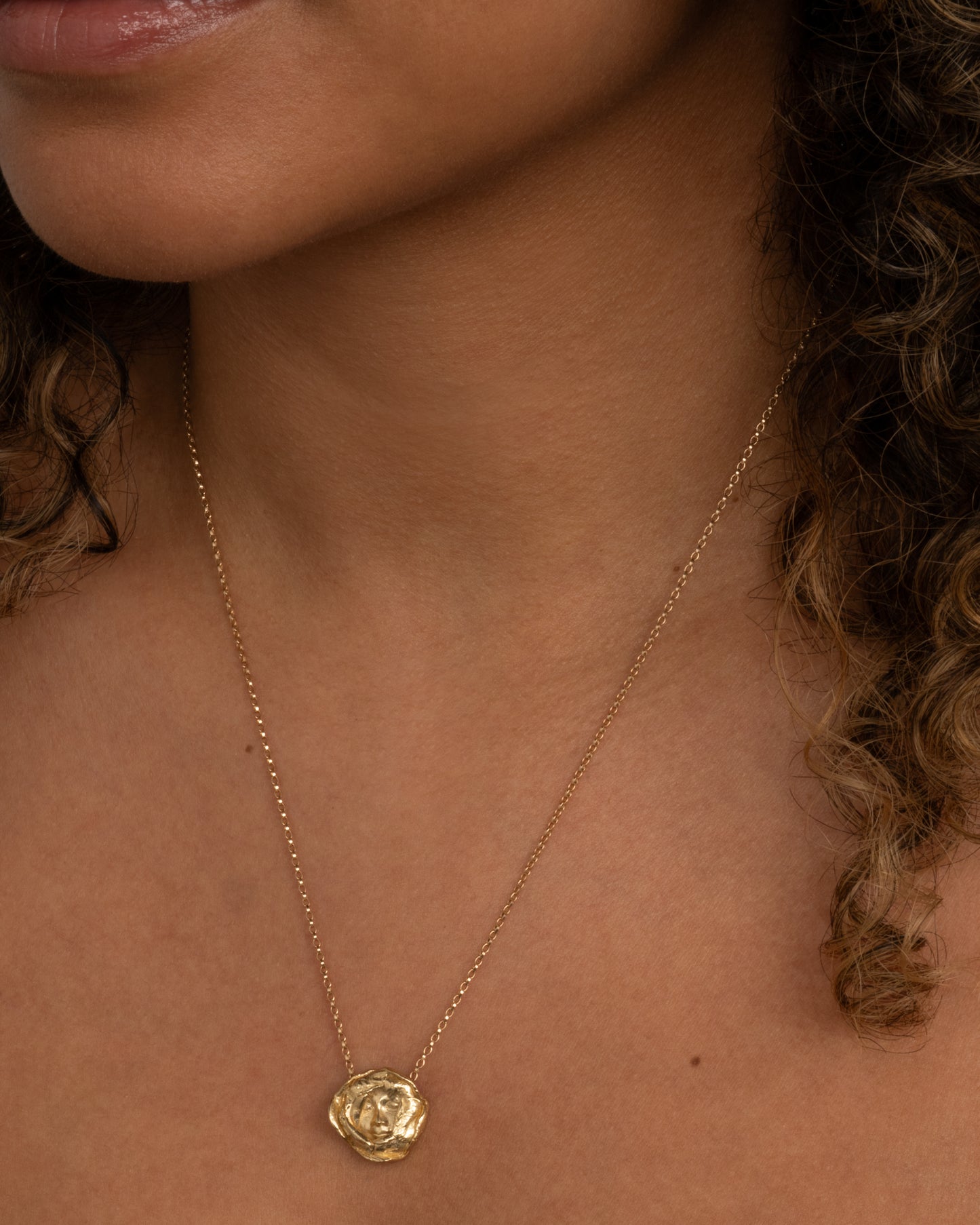 A yellow gold flower pendant with a sleeping face at its center on a cable chain. Shown worn around the neck