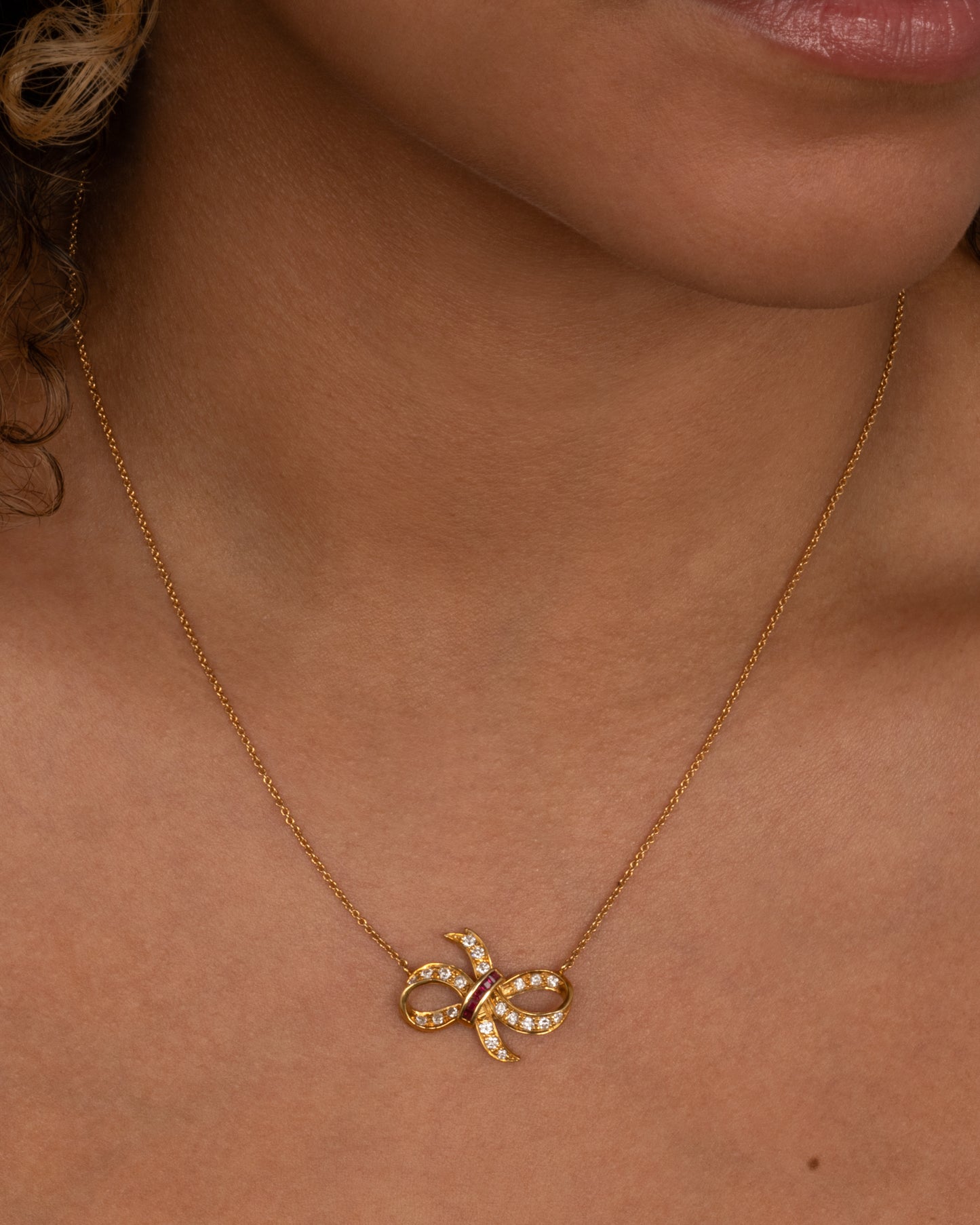 A yellow gold cable chain necklace with a diamond and ruby studded bow at the center. Shown from the front on a person.