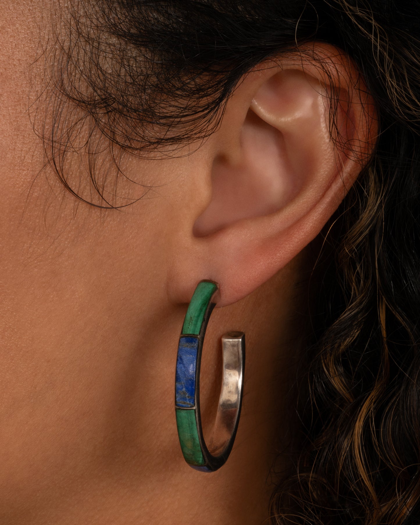 A silver hoop earring with malachite and lapis inlay. Shown on an ear.