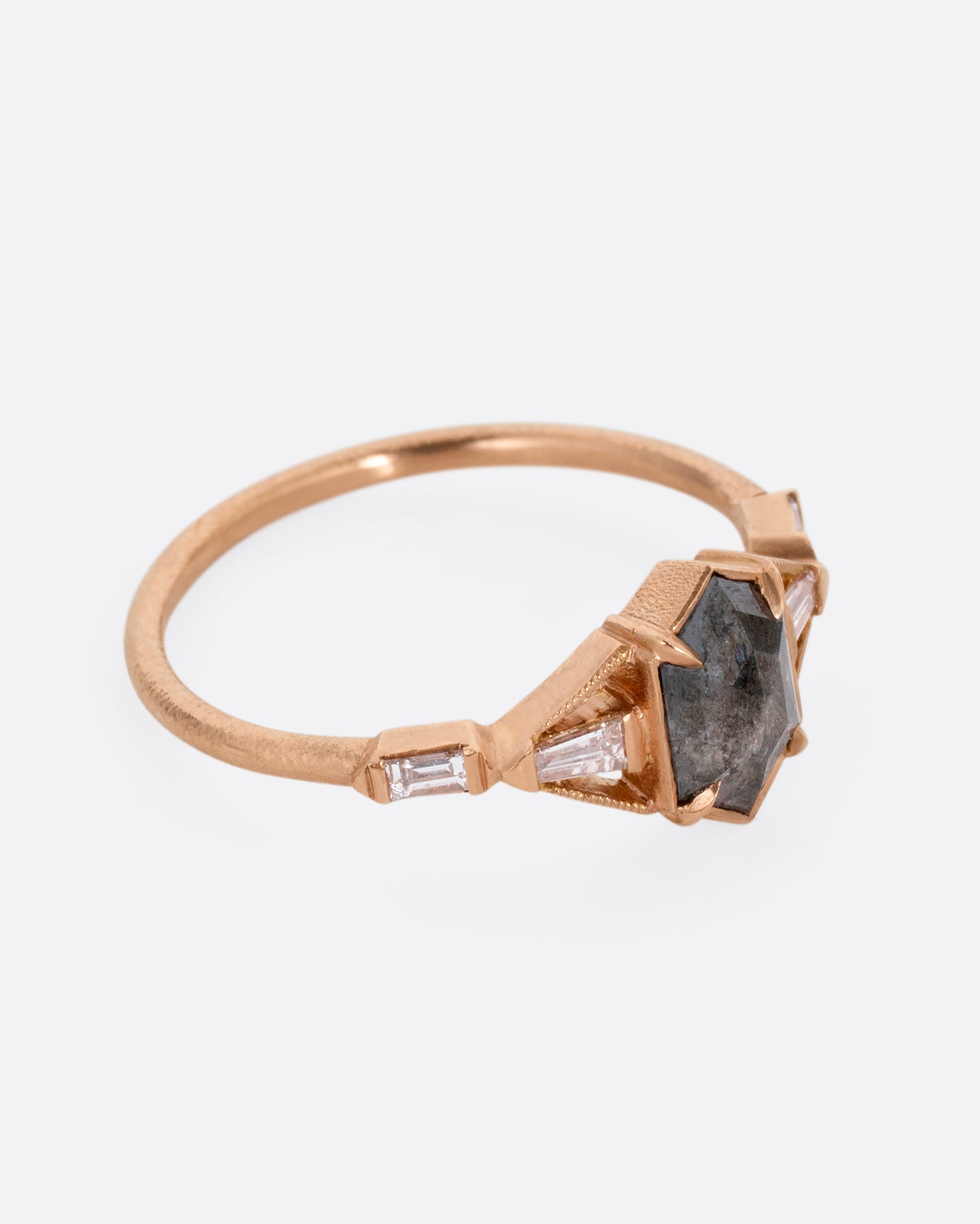 A rose gold ring with a black hexagonal diamond and baguette diamond lined shoulders. Shown from the side.