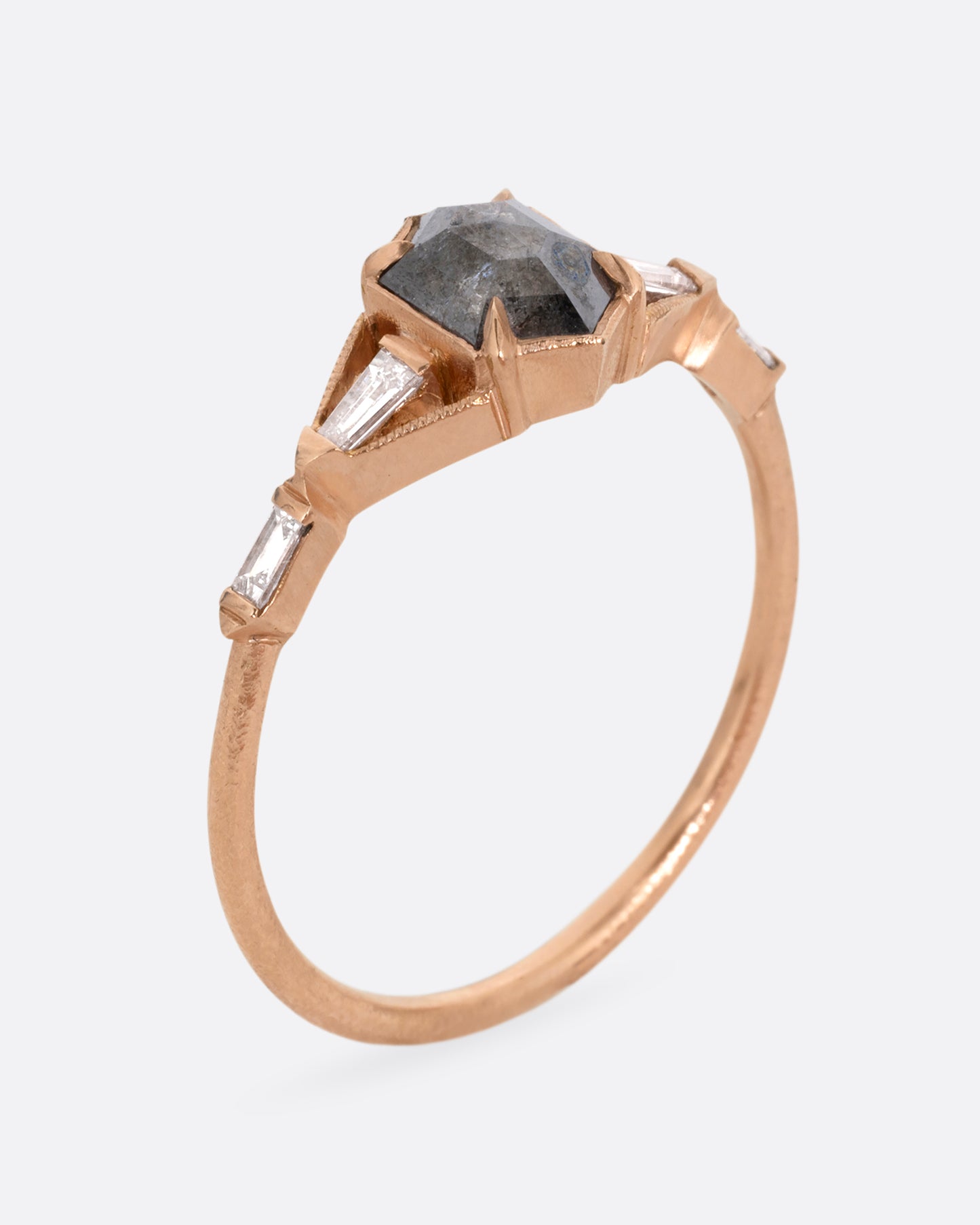 A rose gold ring with a black hexagonal diamond and baguette diamond lined shoulders. Shown standing up from the side.