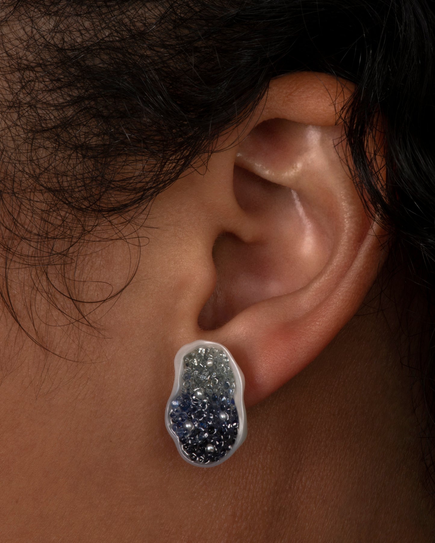 A stud earring made from half of one freshwater soufflé pearl, lined with blue sapphires and seed pearls. Shown on an ear.