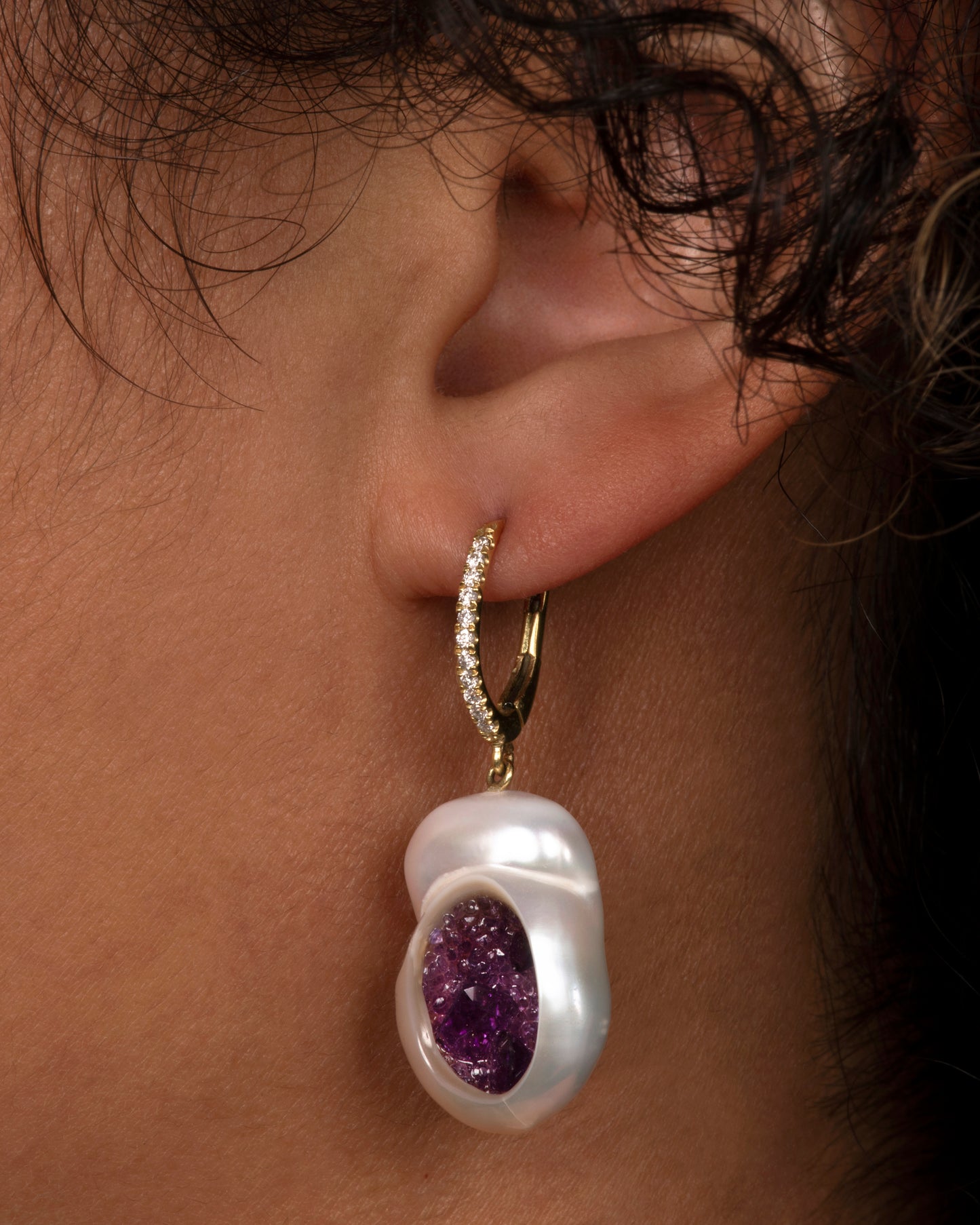 A pair of dangle earrings with carved out pearls lined with amethysts. Each earring is topped with yellow gold ear hook with pave diamonds. Shown from the front on an ear.