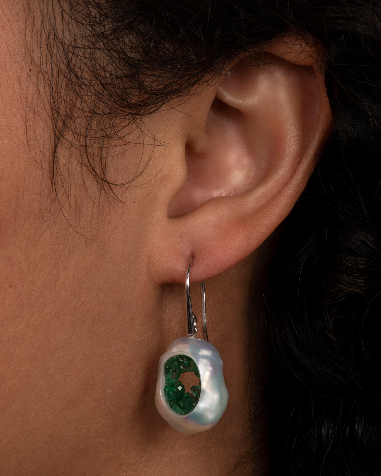 A pair of dangle earrings with hollow pearls lined with emeralds and white gold ear hooks. Shown on an ear.