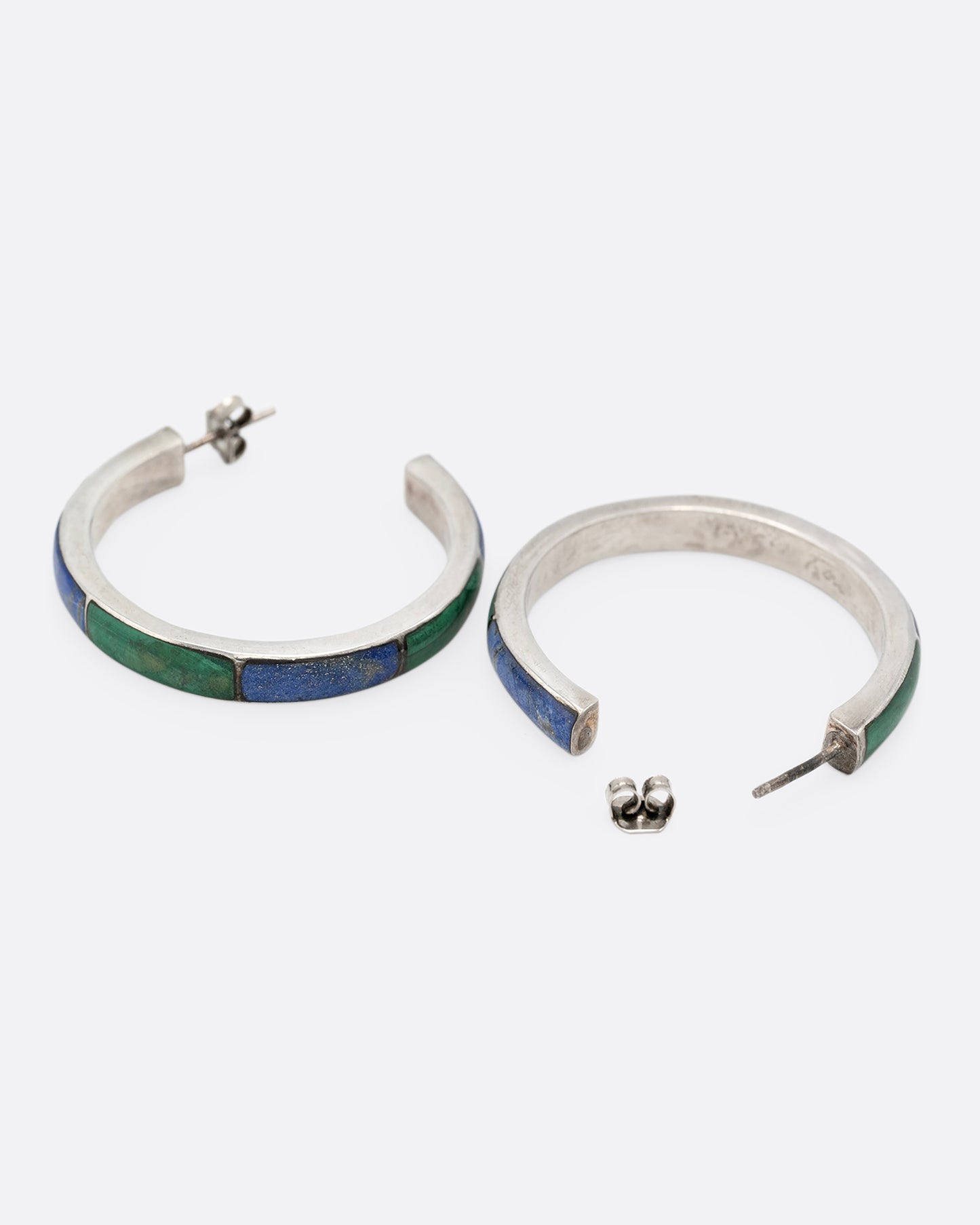 A pair of silver hoop earrings with malachite and lapis inlay. Shown lying on their side with one open.