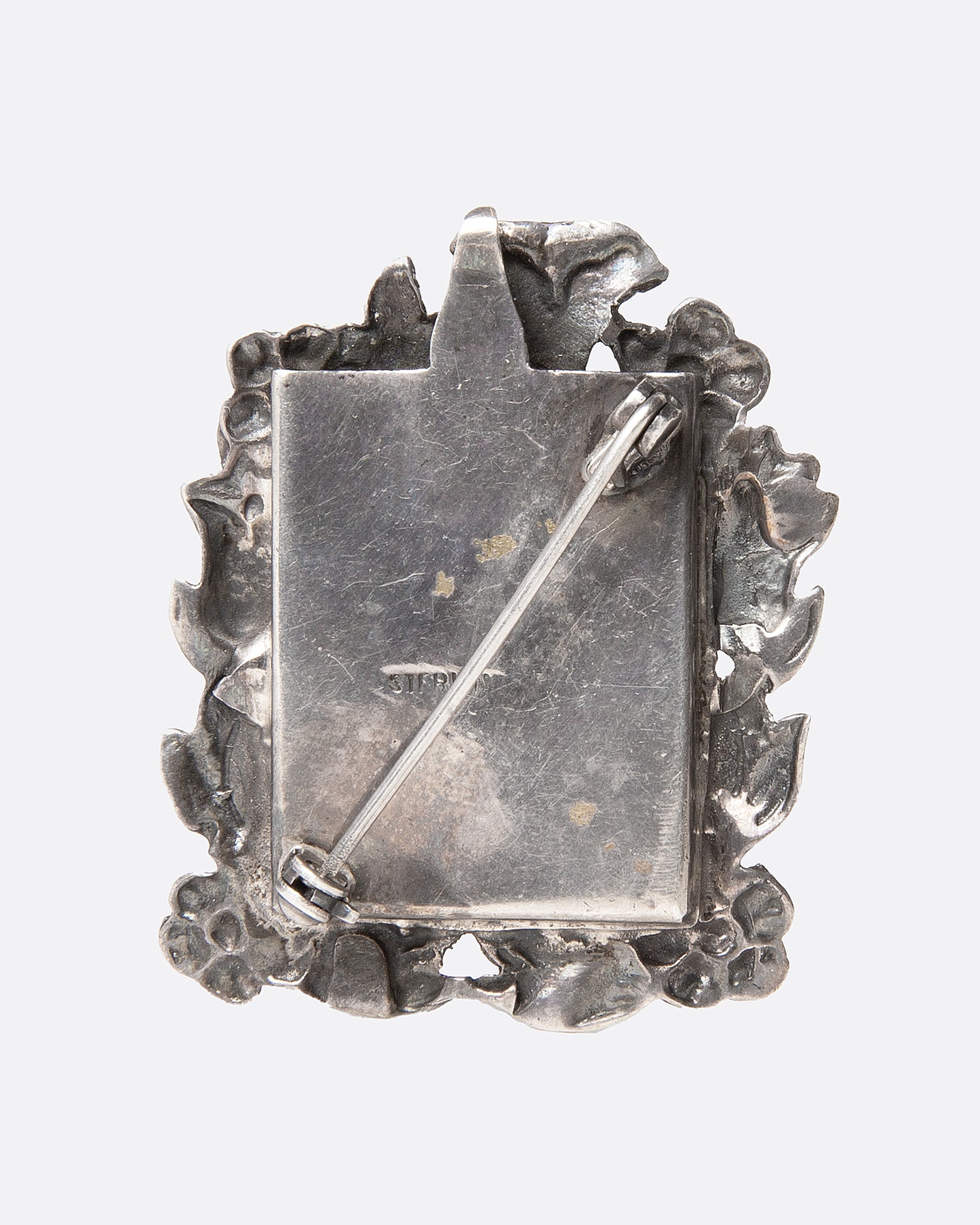An ornate silver picture frame brooch with a botanical motif around a rectangular opening, shown from the back.