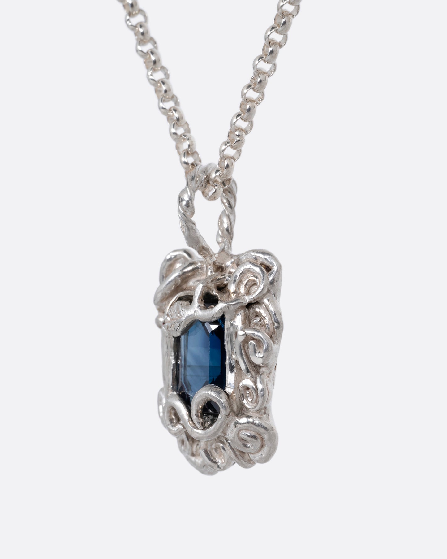 A silver pendant necklace with a deep blue sapphire in a setting with creeping vines. Shown from the side.