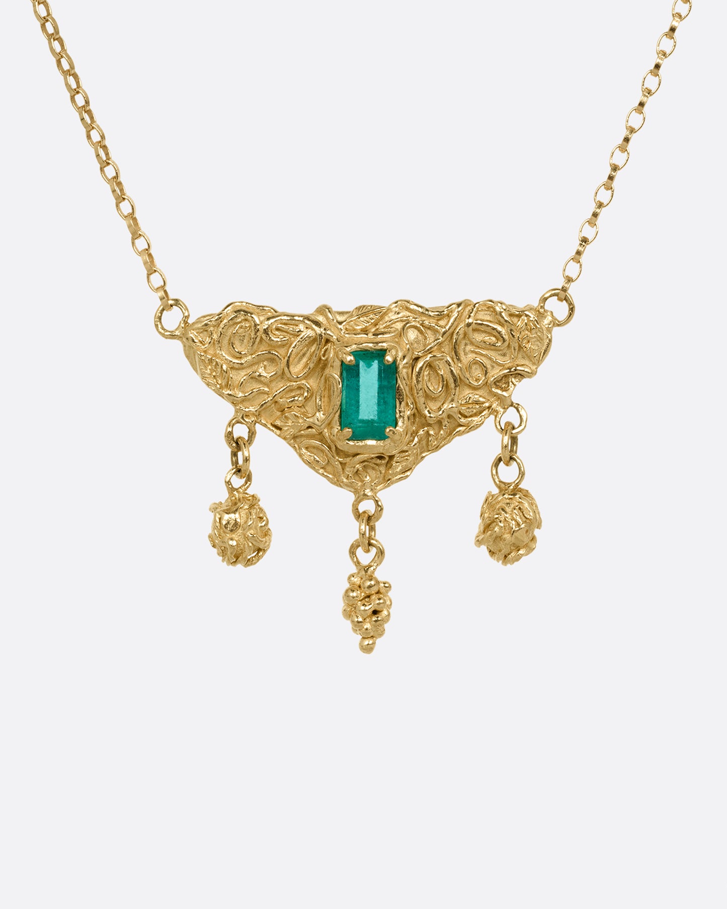 A yellow gold necklace with an emerald at the center of a sculptural pendant and three dangles- two blooming roses and a cluster of grapes. Shown from the front.