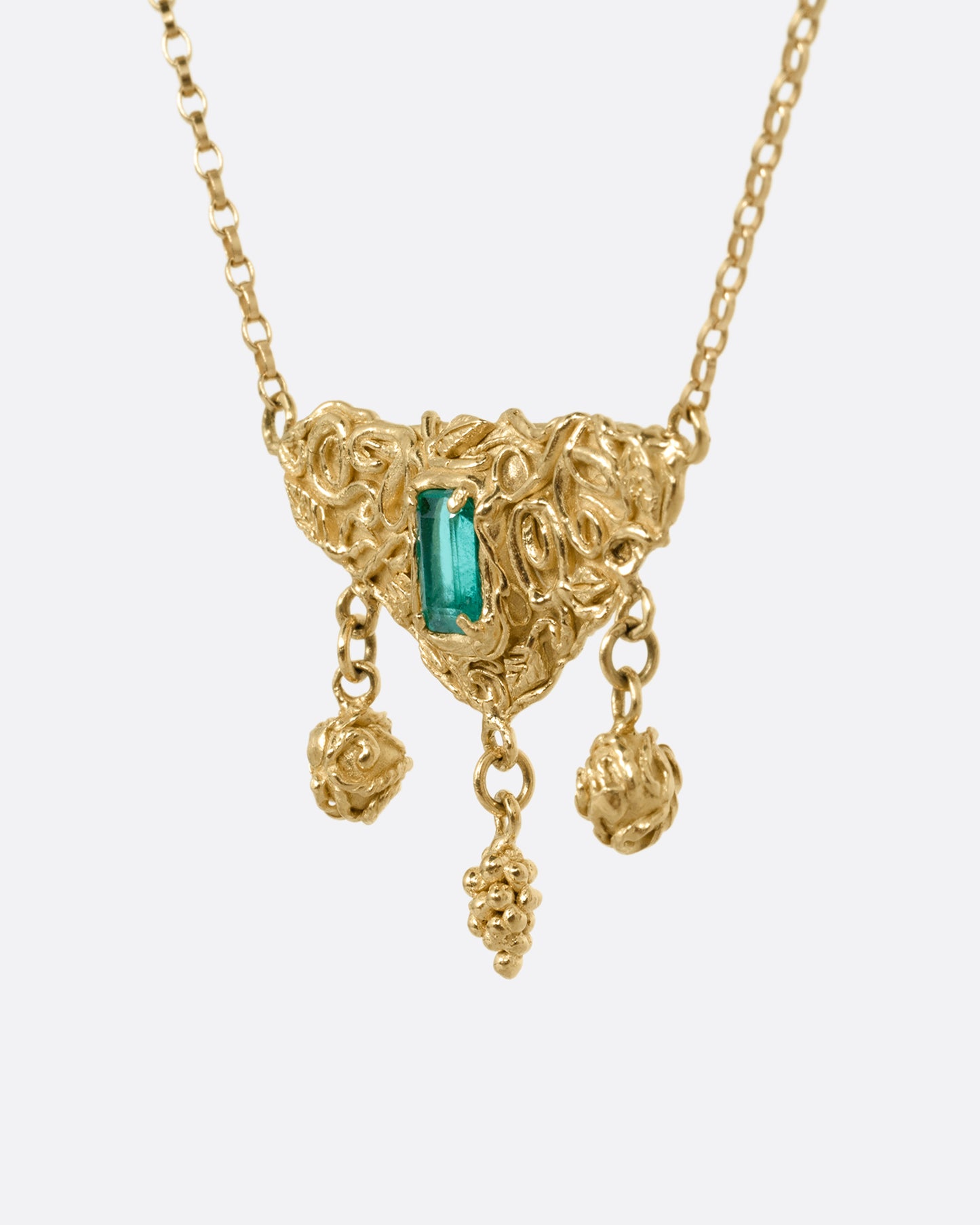 A yellow gold necklace with an emerald at the center of a sculptural pendant and three dangles- two blooming roses and a cluster of grapes. Shown from the side.