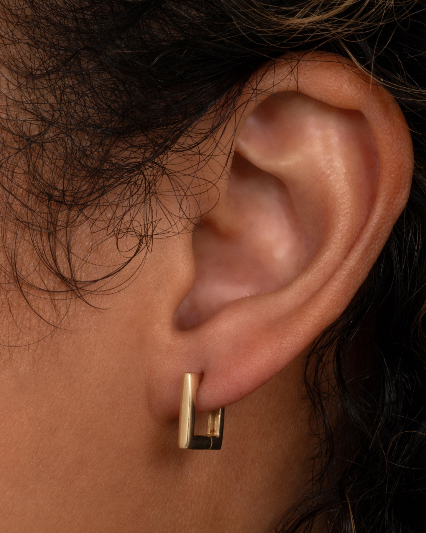 A square yellow gold hoop earring with a hinge clicker clasp. Shown on a person.