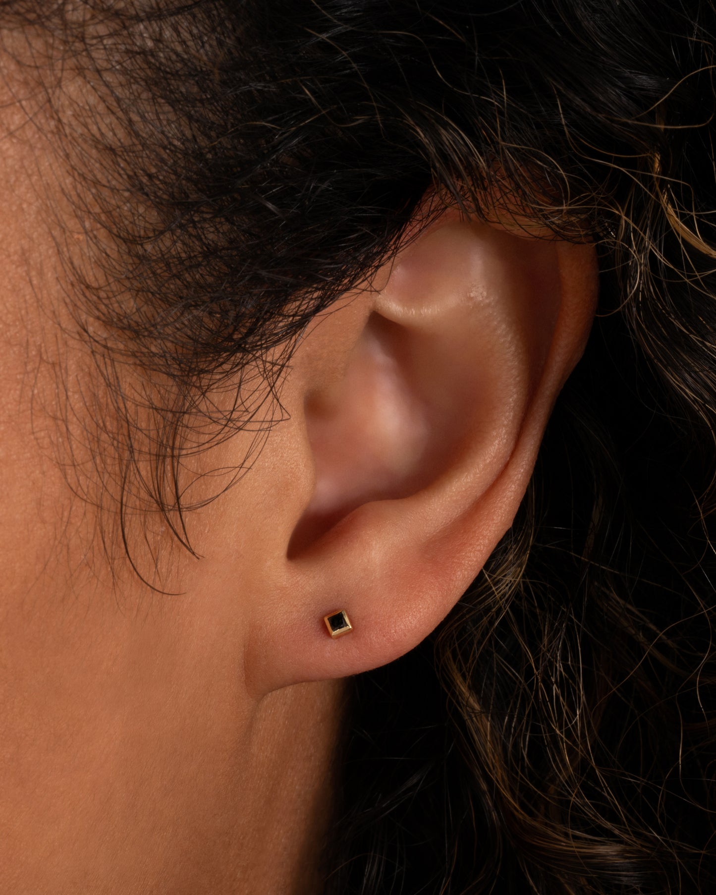 A yellow gold stud earring with a princess cut black diamond. Shown in an ear.