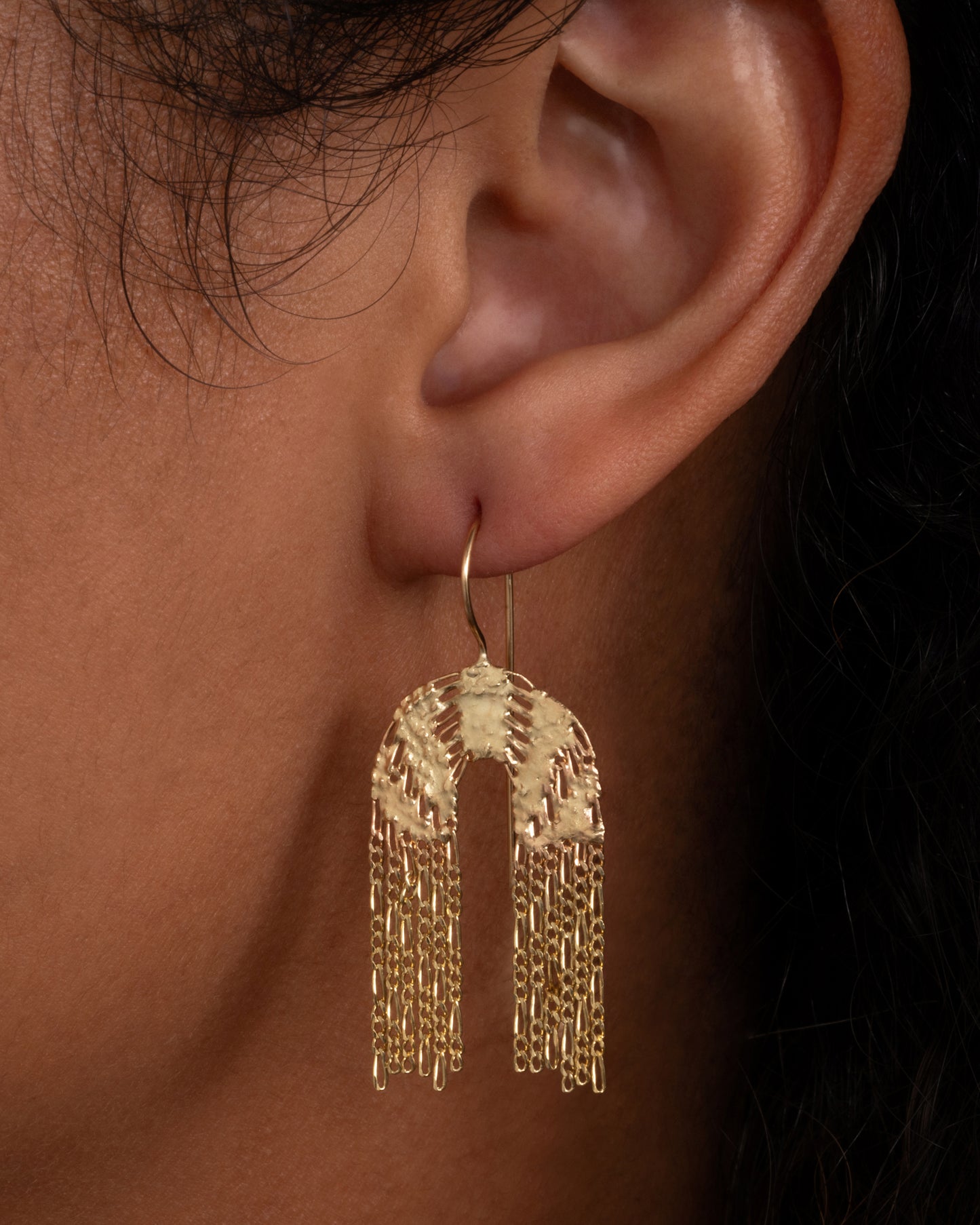 A pair of yellow gold dangle earrings with soldered rainbow shaped tops and Figaro chain fringe bottoms. Shown worn in an ear.