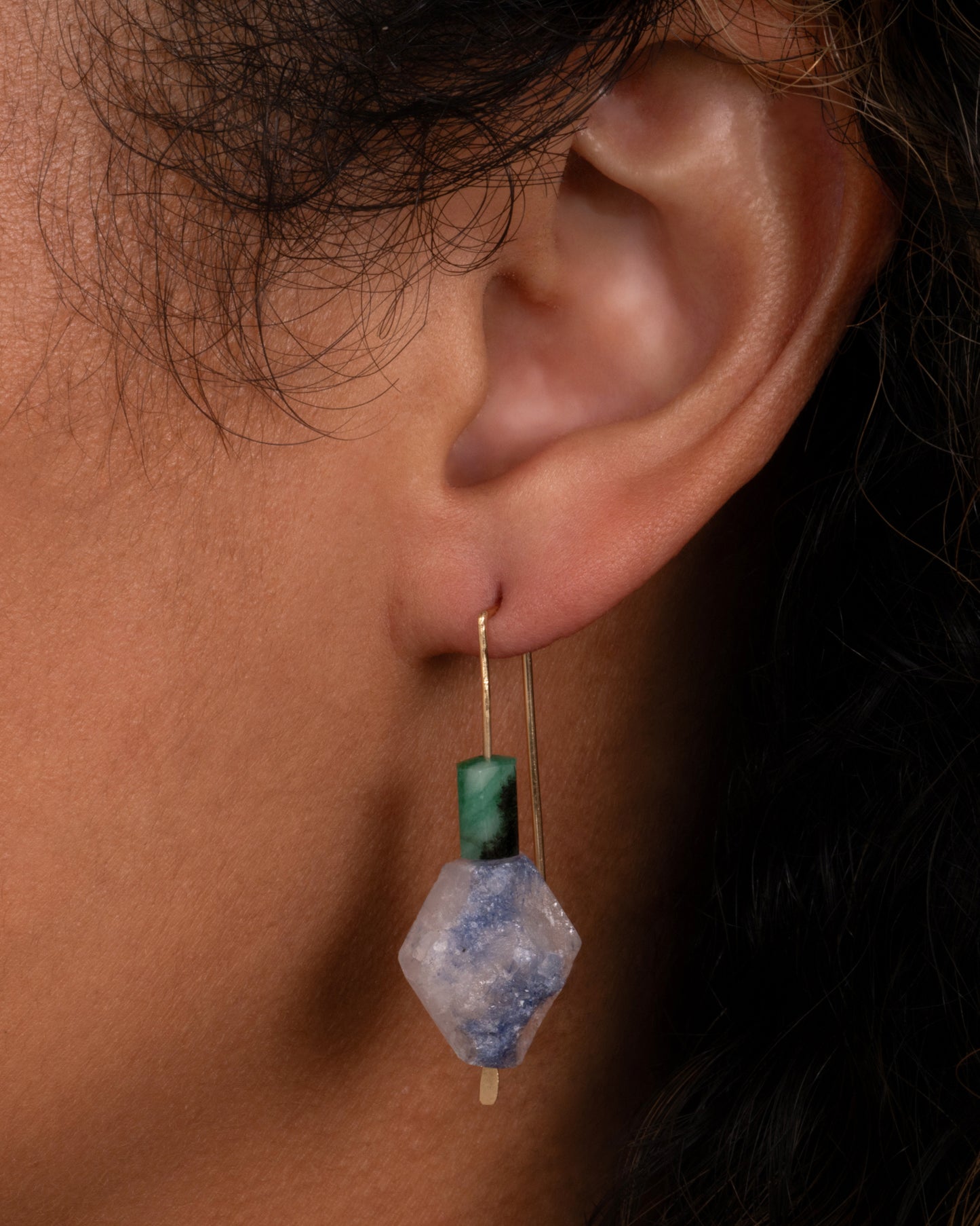 A yellow gold hook earring with a freeform dumortierite and emerald. Shown on an ear.