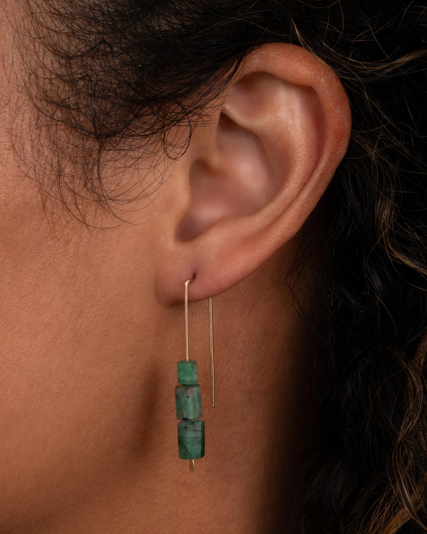 A yellow gold hook earring with three stacked freeform emeralds. Shown on an ear.