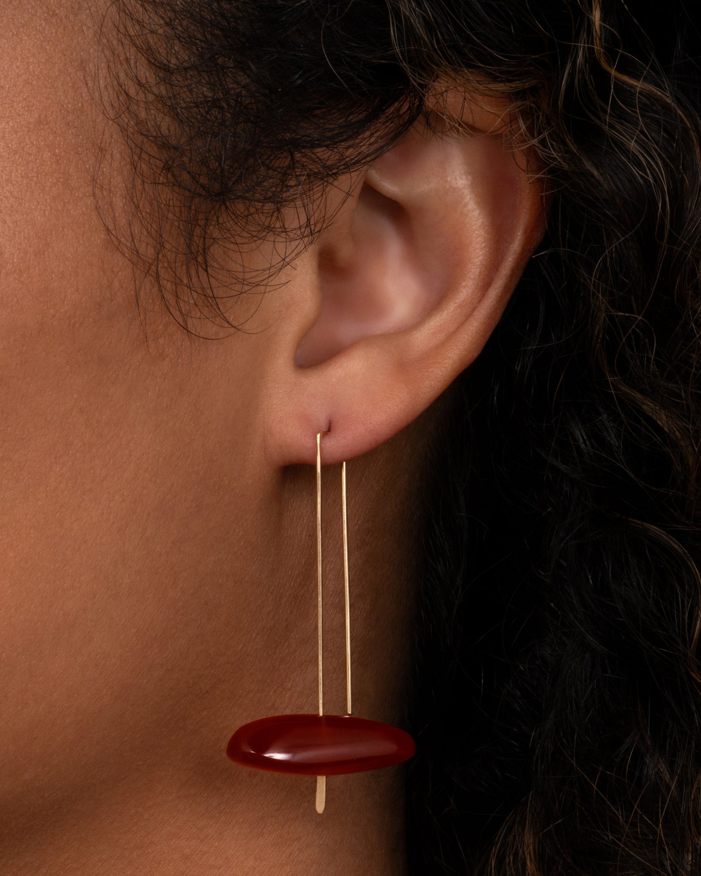 A long yellow gold earring with a freeform carnelian at the base, shown on an ear.