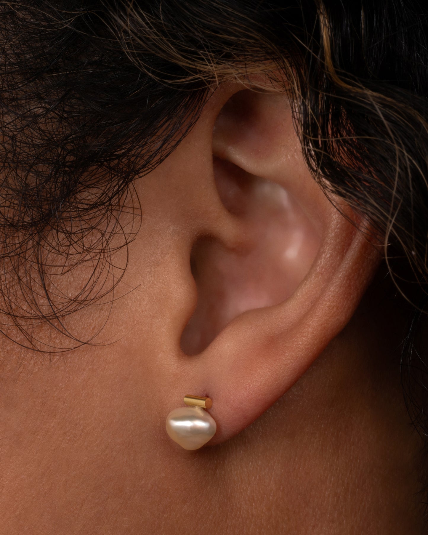 A pearl earring suspended from a gold bar setting. Shown worn in an ear.