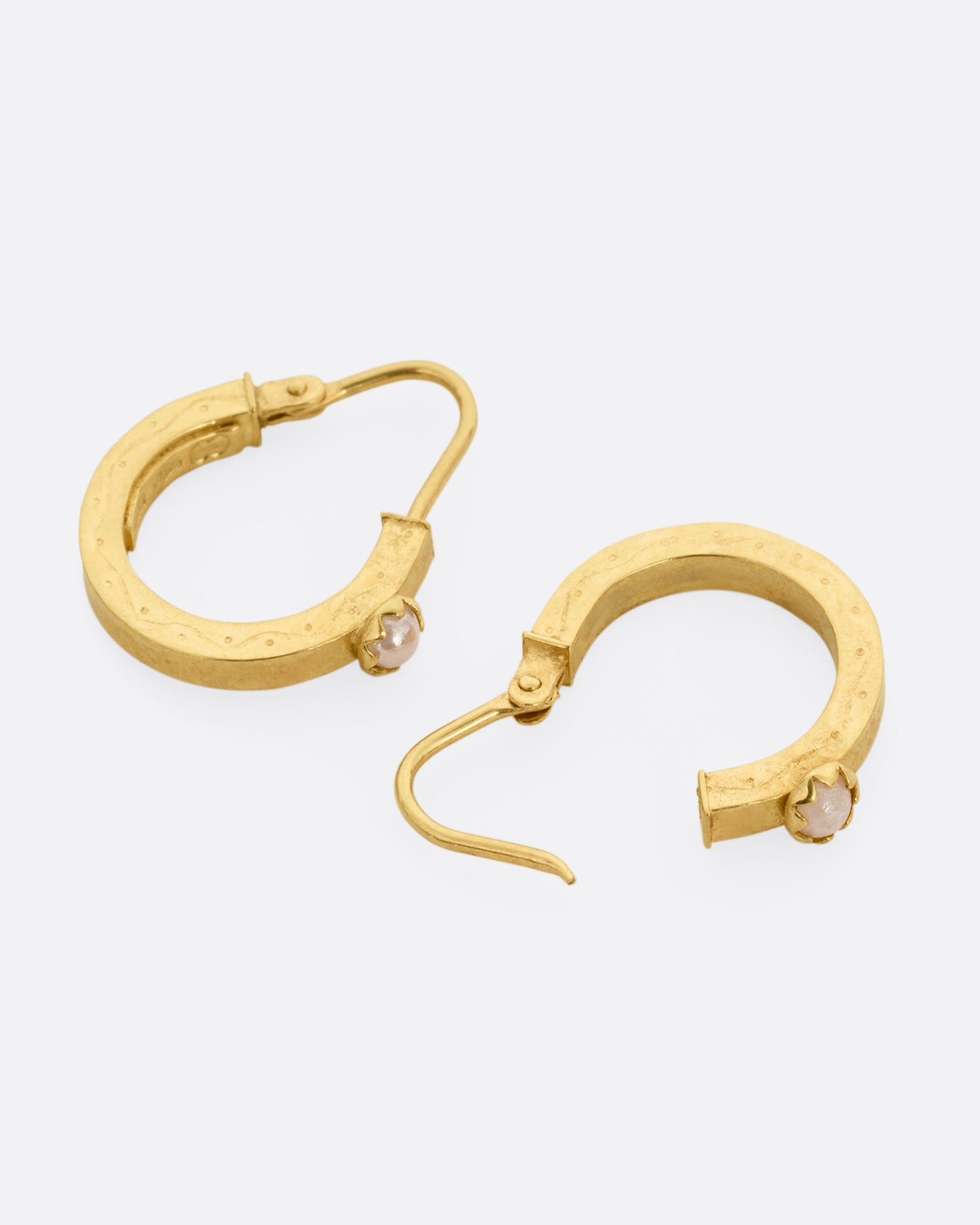 A pair of yellow gold hoop earrings, each with a pearl, shown lying flat with one earring open.