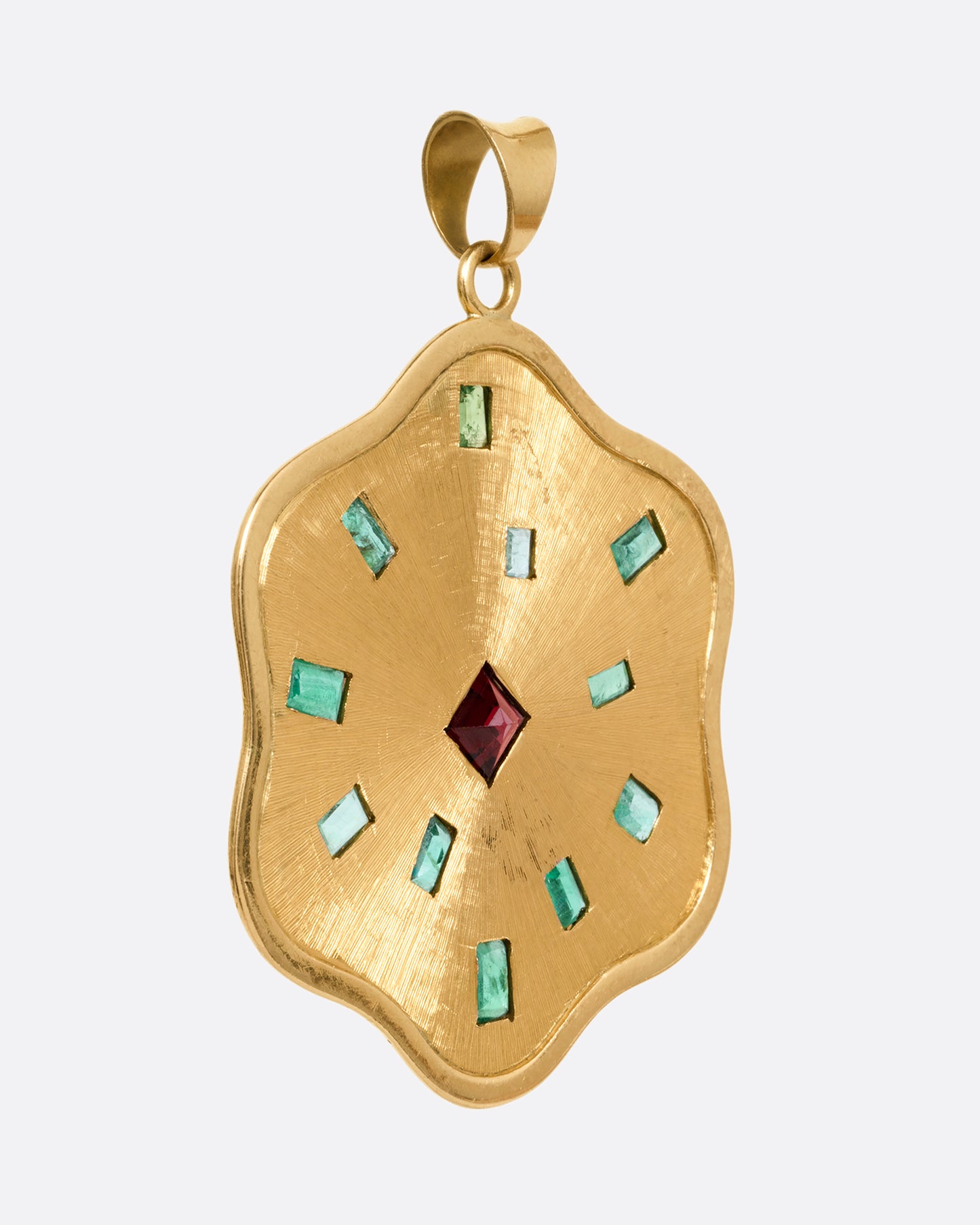 A yellow gold wavy edged medallion pendant with baguette emeralds encircling a central princess cut garnet. Shown from the side.