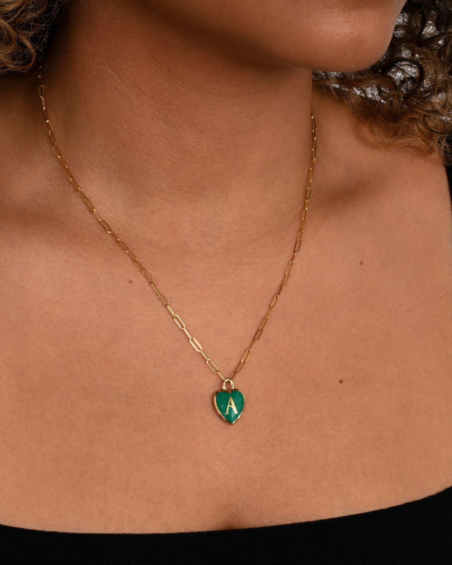 A yellow gold paperclip chain necklace with a green enamel heart shaped A pendant. Shown from the front on a person.