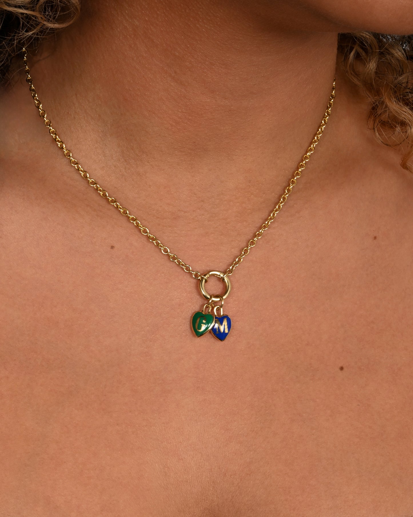 A yellow gold cable chain necklace with a ring at the center. A blue enamel heart with a letter M and a green enamel heart with a letter G hang from the center ring. Shown from the front on a person.