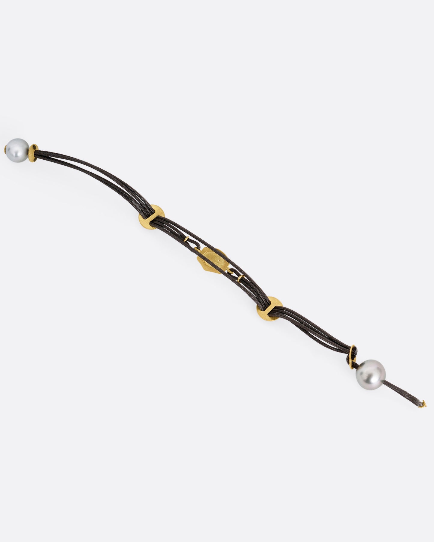 A black cord bracelet with a diamond studded tag flanked by two gold slides and a pearl closure. Shown open from the back.