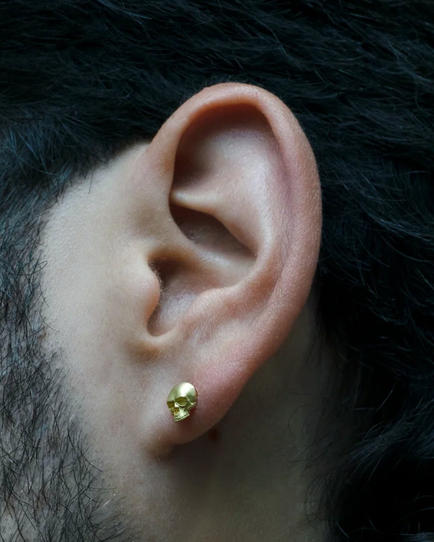 A solid yellow gold skull stud earring. Shown on an ear from the front.