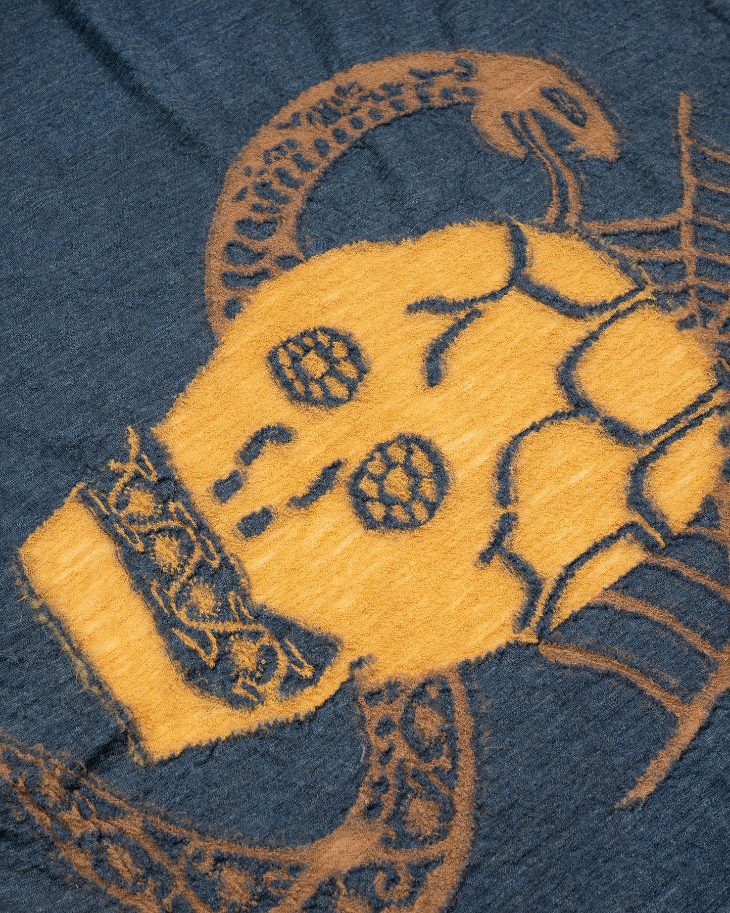 A navy blue and grey scarf with a golden yellow skull in the center. View close up of the skull.