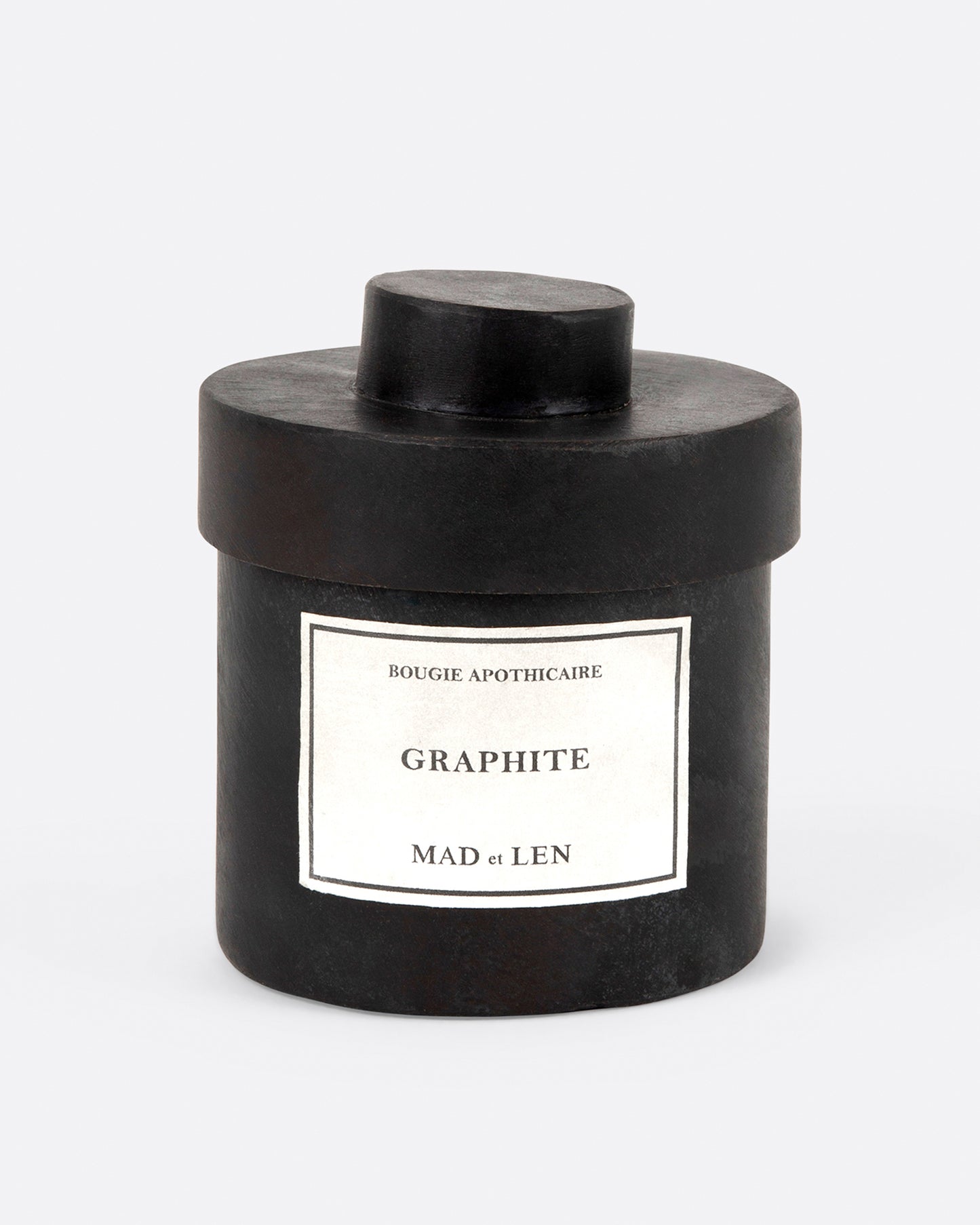 A Mad et Len bois Graphite candle measuring 3" tall and 3.25" wide. It is housed in a one-of-a-kind blackened iron vessel with a lid and a label that reads: Bougie Apothicaire Graphite Mad et Len. Shown from the front.