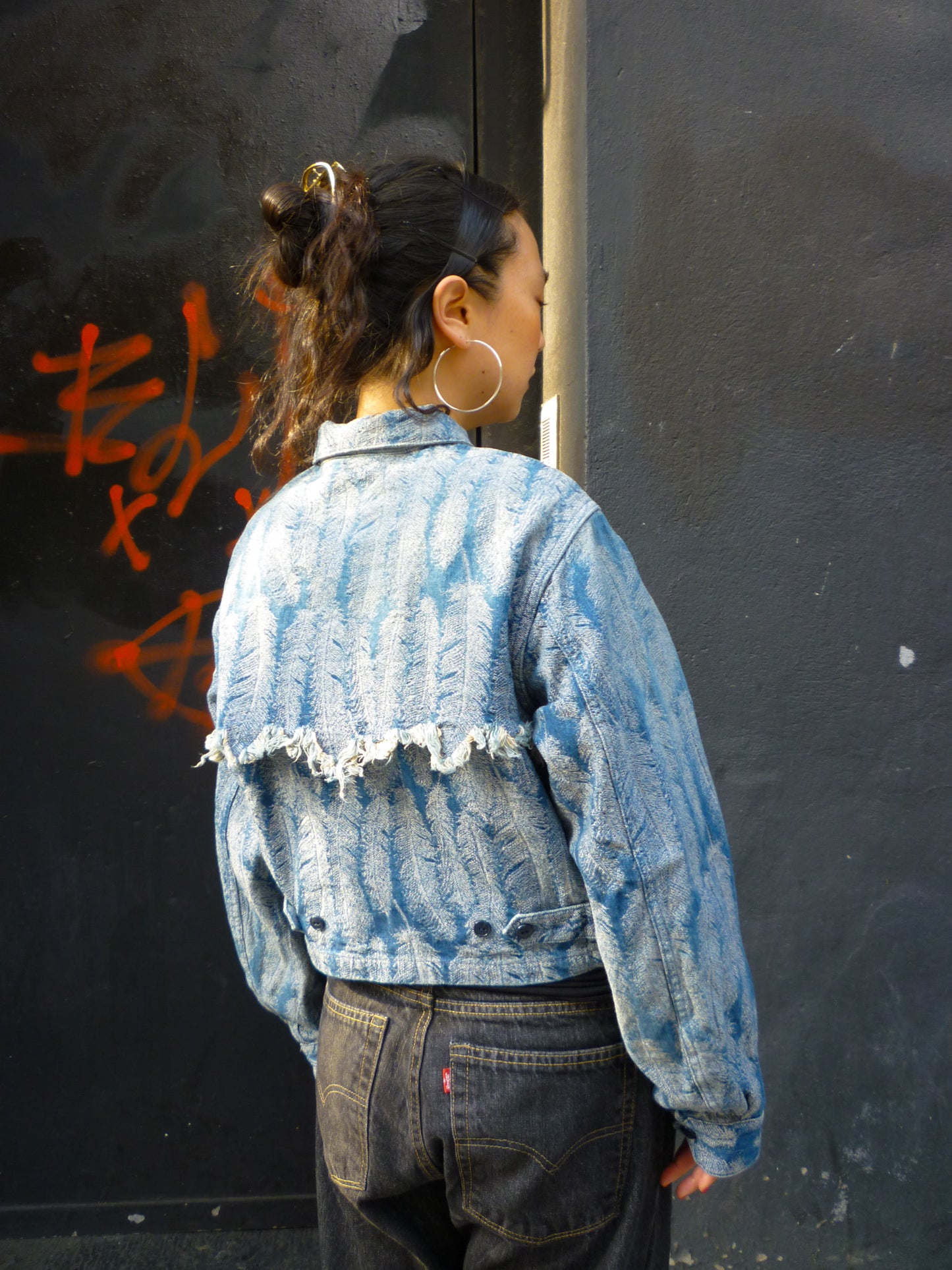 A light blue jacquard denim jacket with an all- over feather motif, and a cut-off hemline with raw feathered seam. Shown from the back on a person.