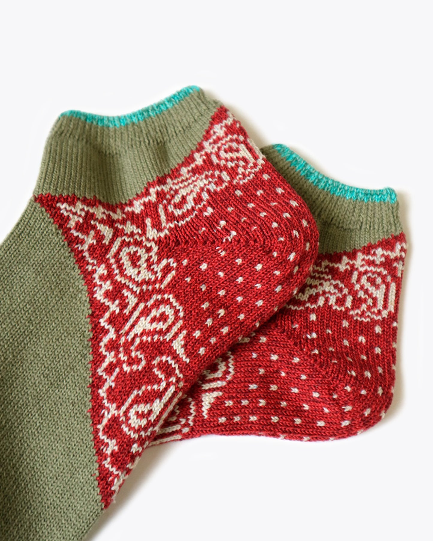 A close up of the heels of a pair of sage green and turquoise green socks with a red paisley bandana print heel. Shown lying flat from the side.