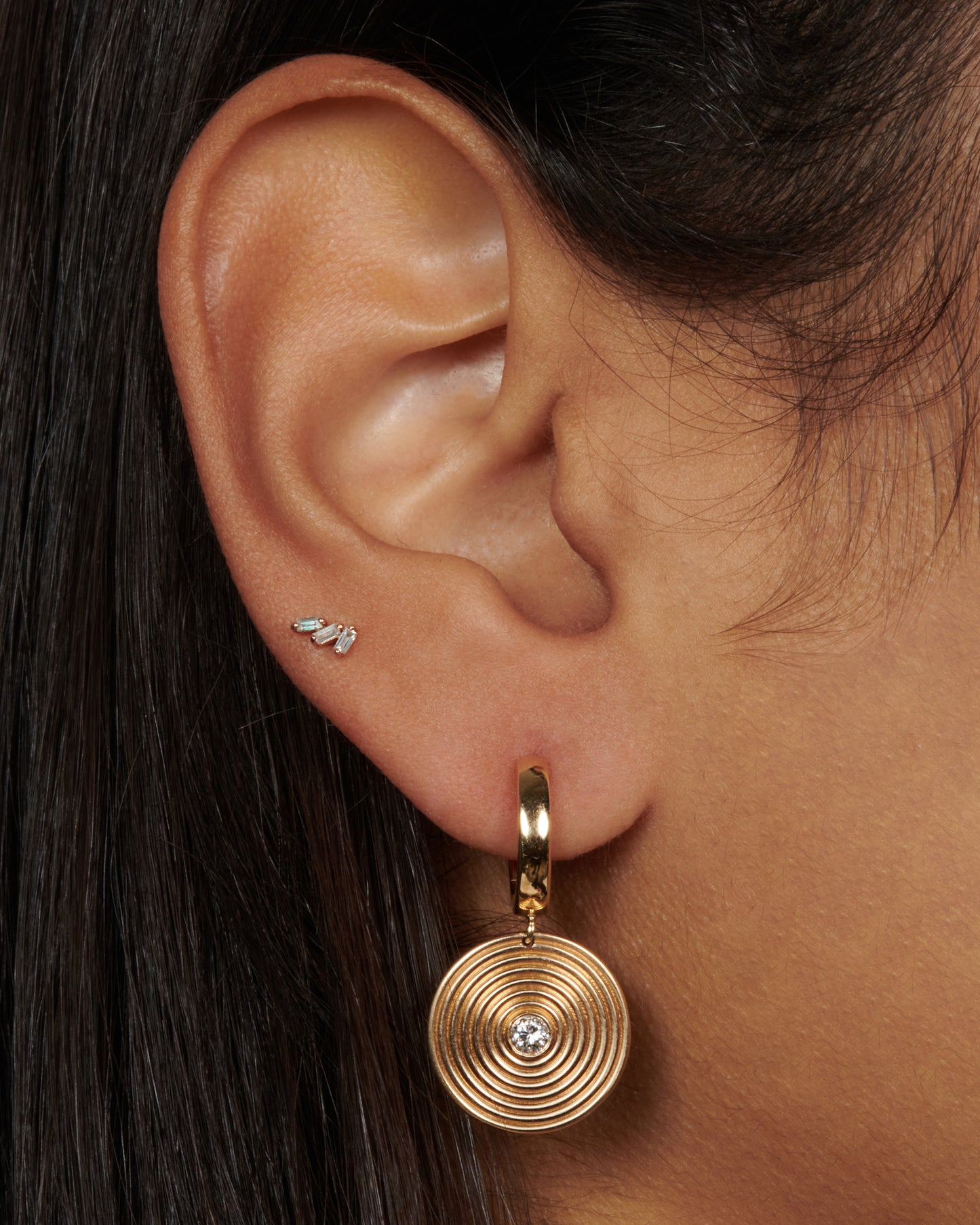 A yellow gold hoop earring with a circular ribbed disc drop with a white diamond center, shown from the front on a person.
