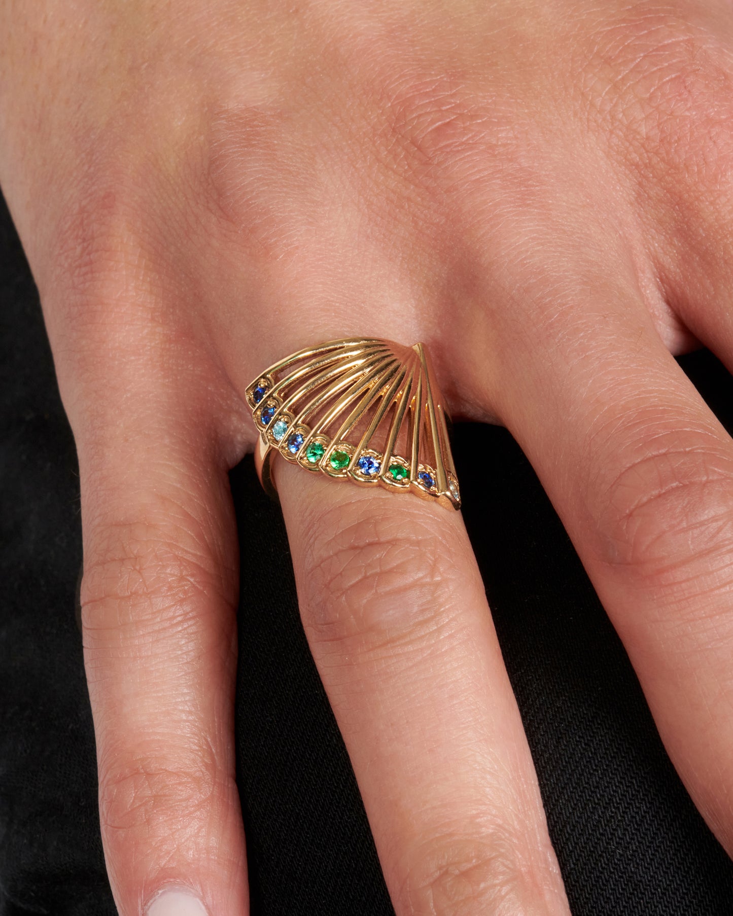 A yellow gold ring in the shape of a fan with multicolored sapphires along the top edge. Shown from the front on a hand.