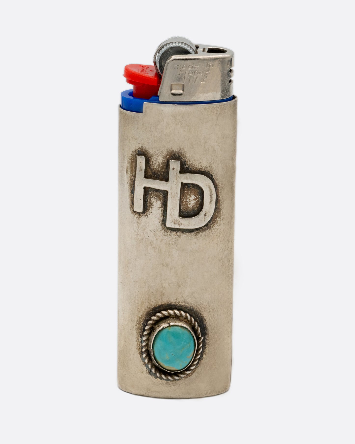 A silver lighter cover with the initials 'HD' and a circular turquoise. Shown from the side.