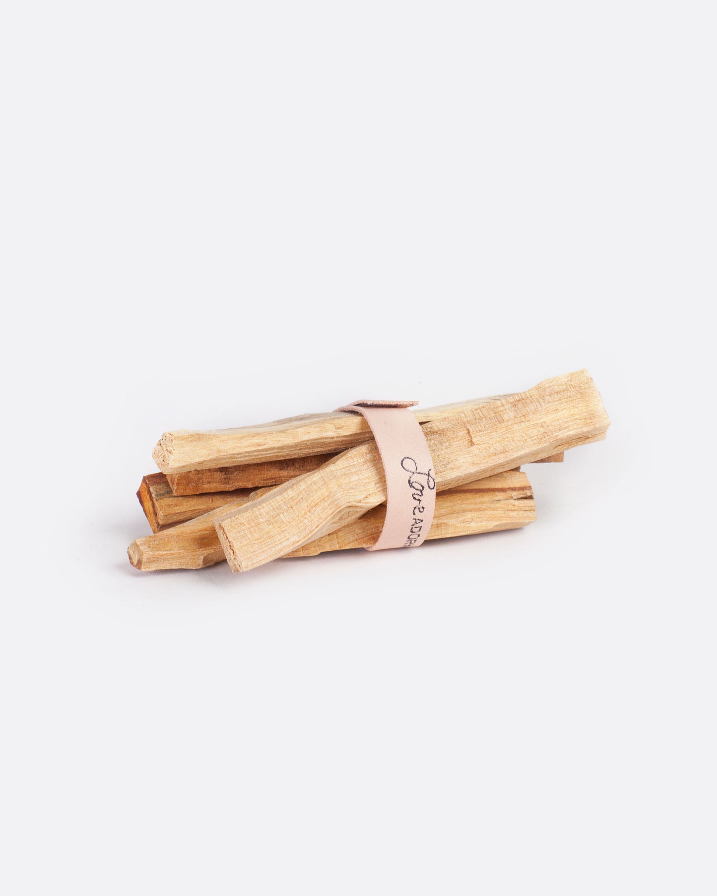 A bundle of a few 4" x 1/2" chunks of palo santo wrapped neatly together with a piece of peach leather that's been stamped with a love adorned logo. Shown from the side.
