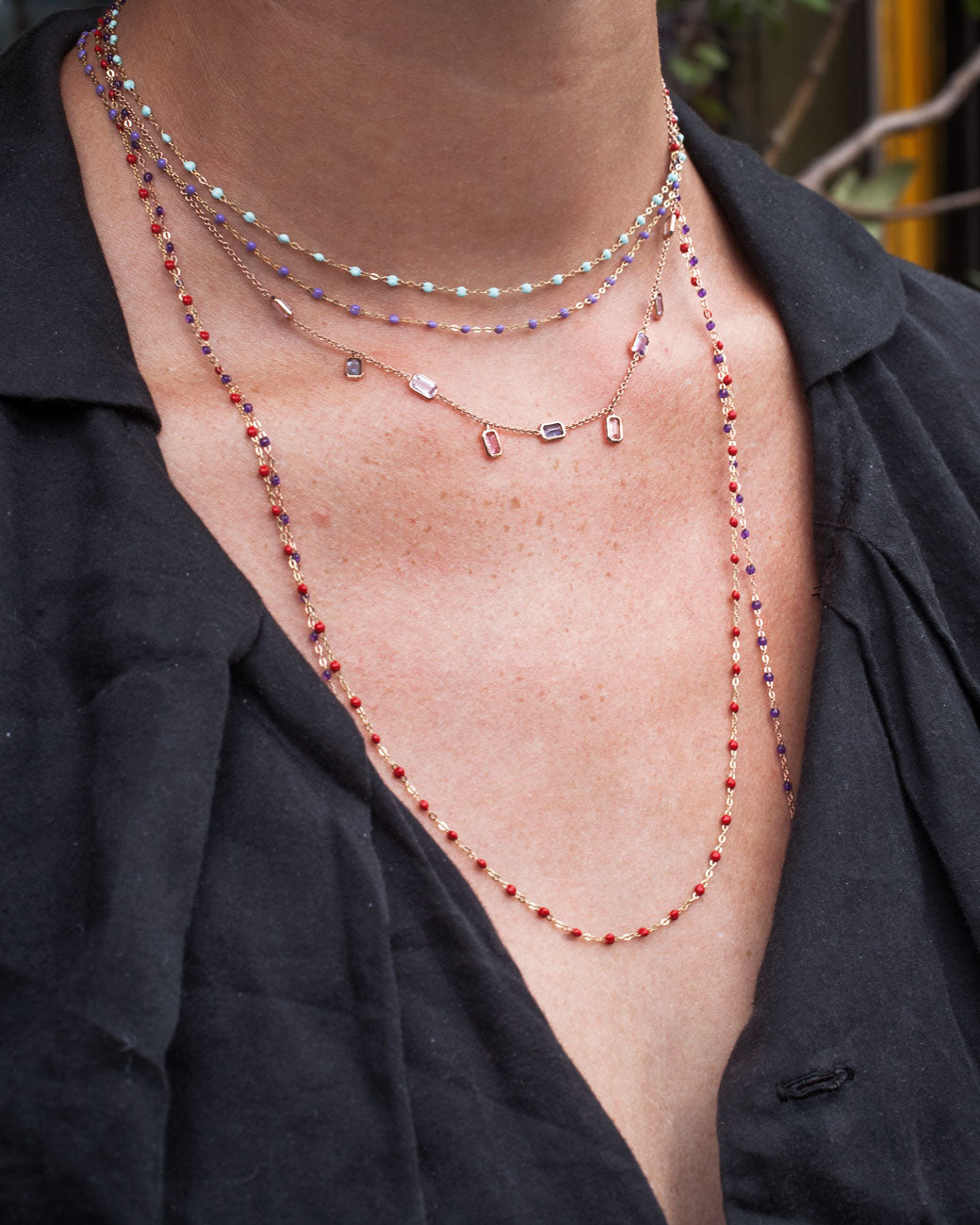 Closeup of woman's neck; she wears multiple yellow gold chain necklaces with resin beads of various lengths in addition to another necklace with precious gemstones and a black shirt. Shown from the front.