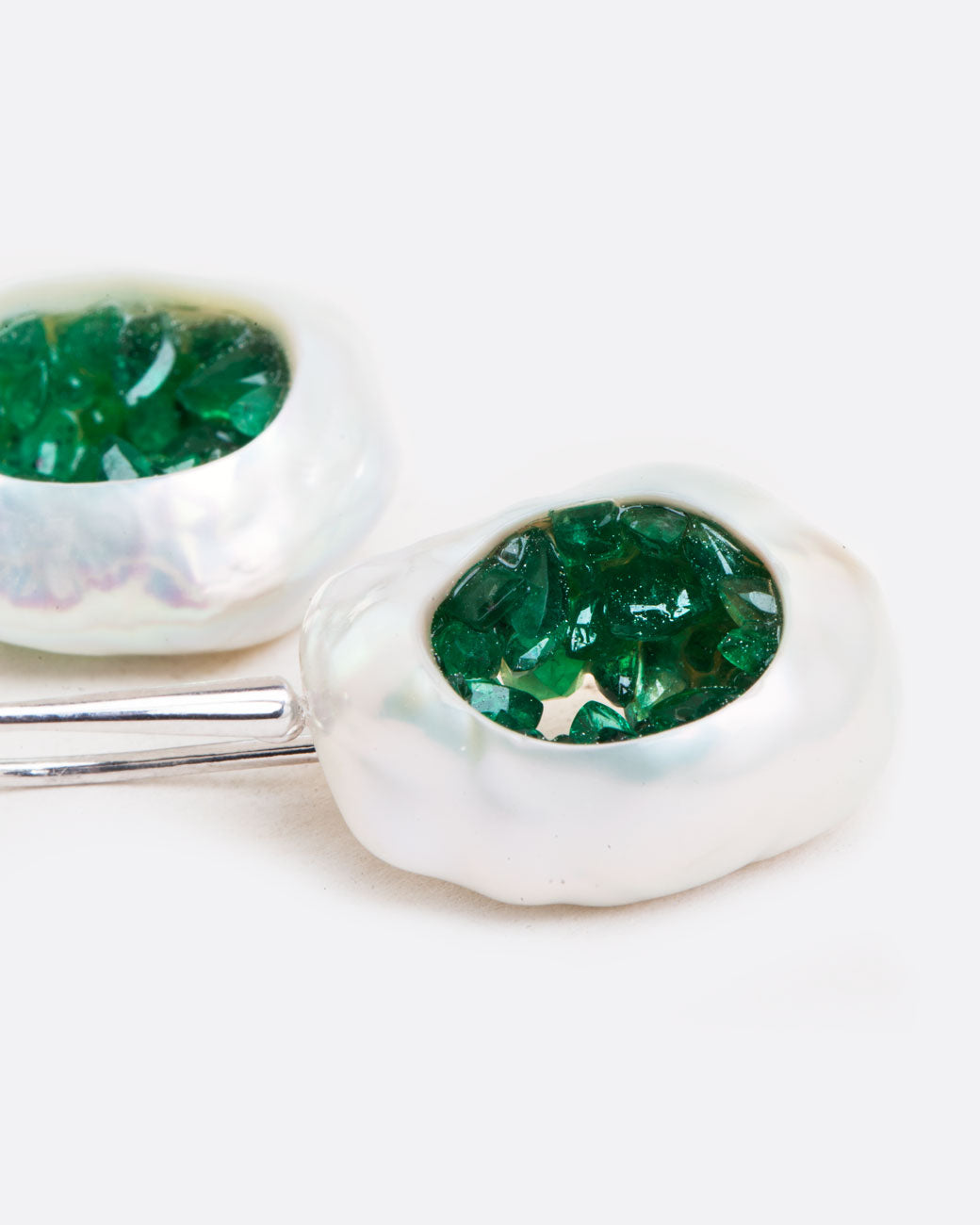 A pair of dangle earrings with hollow pearls lined with emeralds and white gold ear hooks. Shown lying flat from the side.