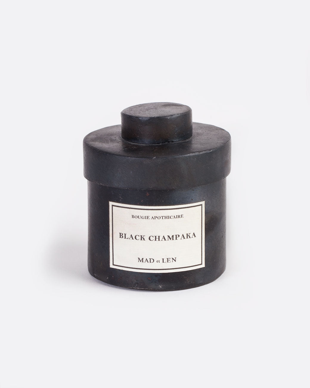 A Mad et Len Black Champaka candle measuring 3" tall and 3.25" wide. It is housed in a blackened iron vessel with lid and a label that reads: Bougie Apothicaire Black Champaka Mad et Len. Shown from the front.